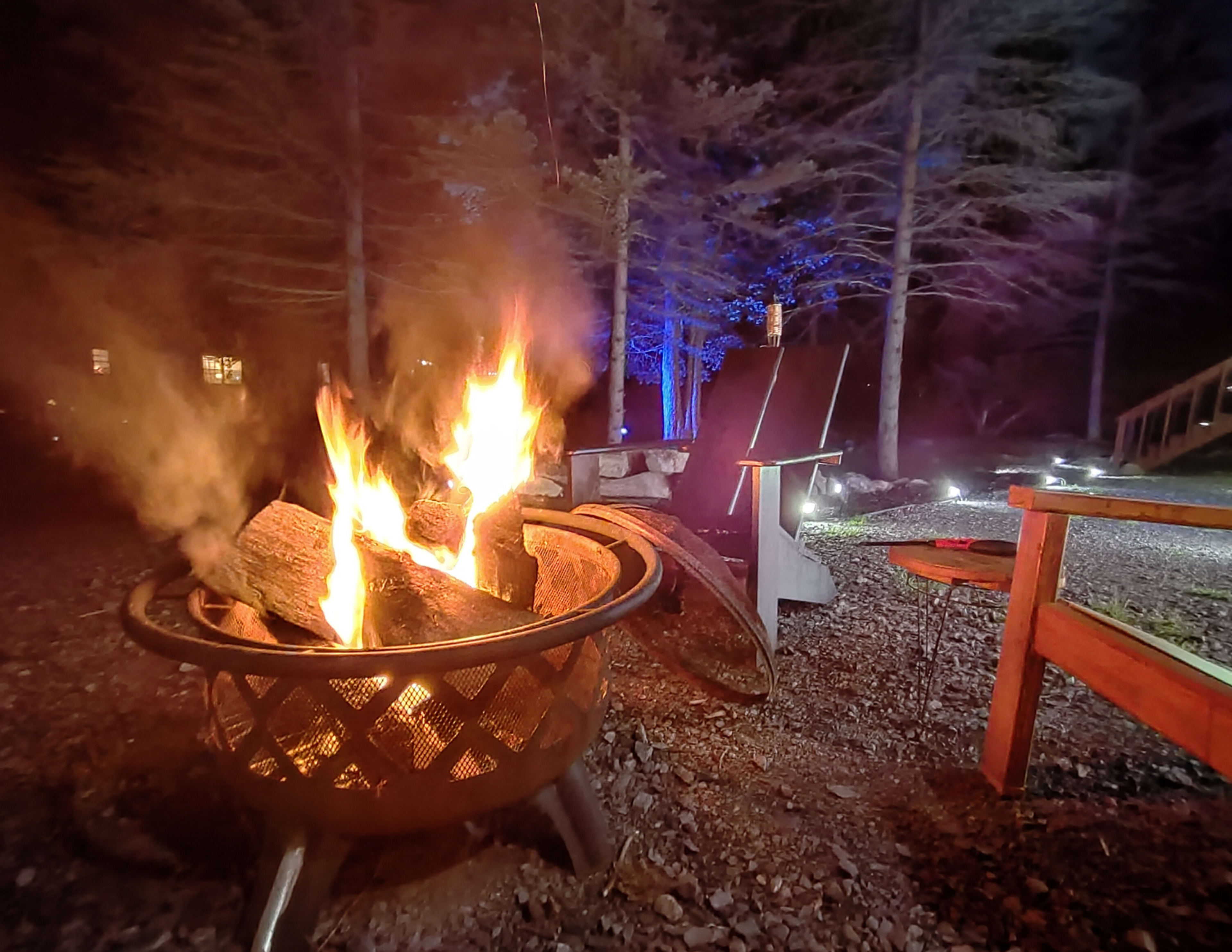 A brightly burning campfire flickers in a fire pit surrounded by wooden chairs in a wooded area at night.
