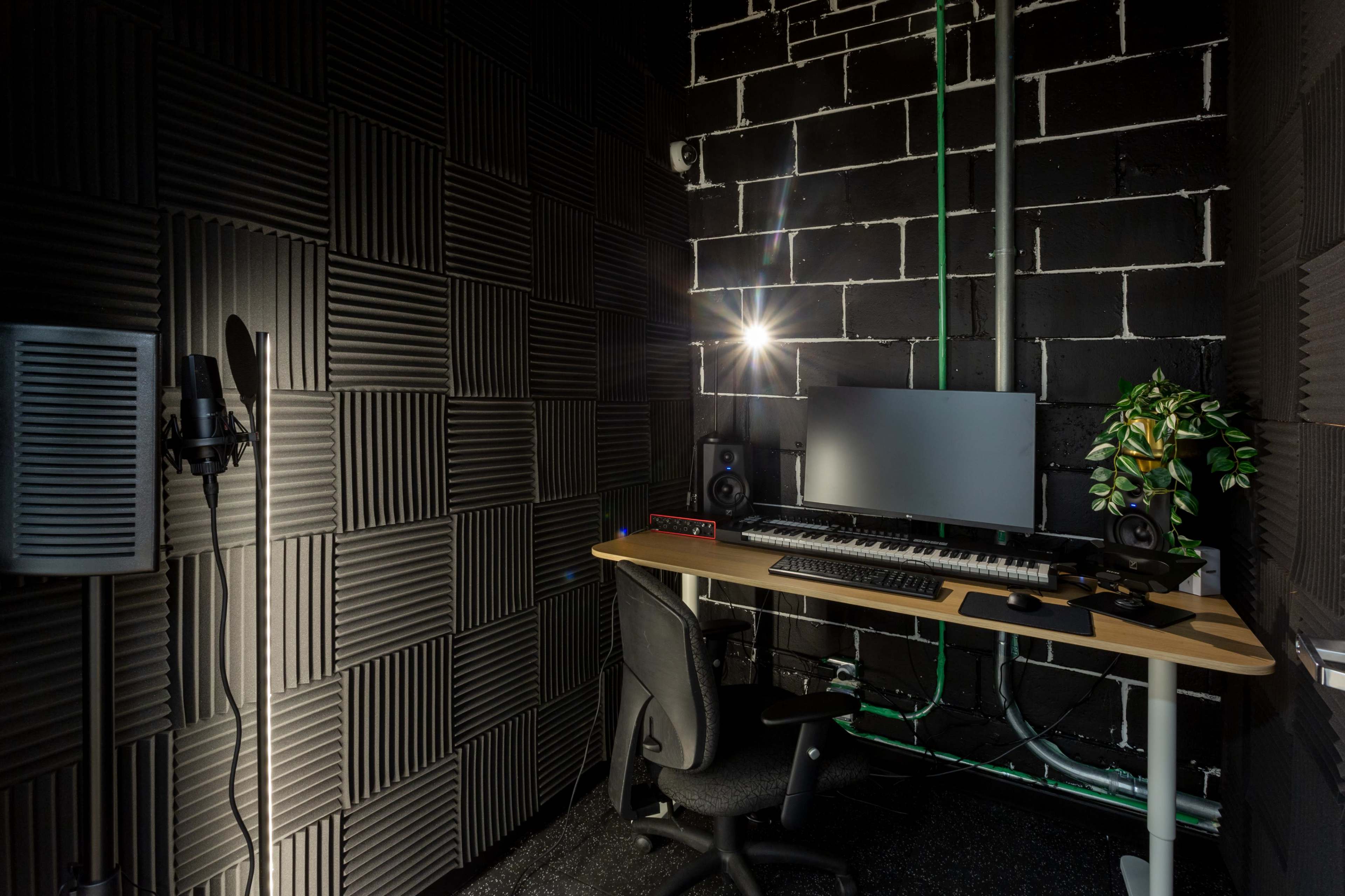 The image shows a soundproofed recording studio featuring a desk with a computer, keyboard, monitor, and speakers, along with a microphone and a plant in the corner.