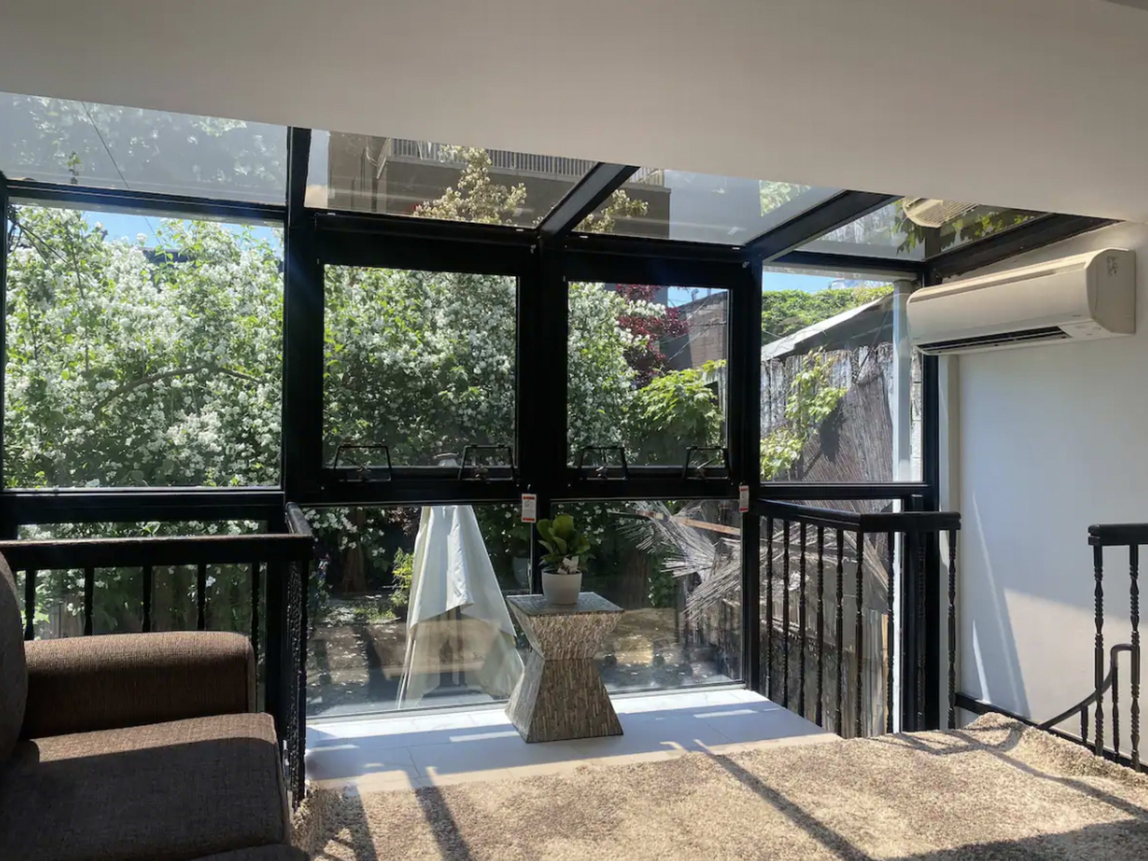 The image shows a modern living space with large glass windows that open to a garden filled with greenery.