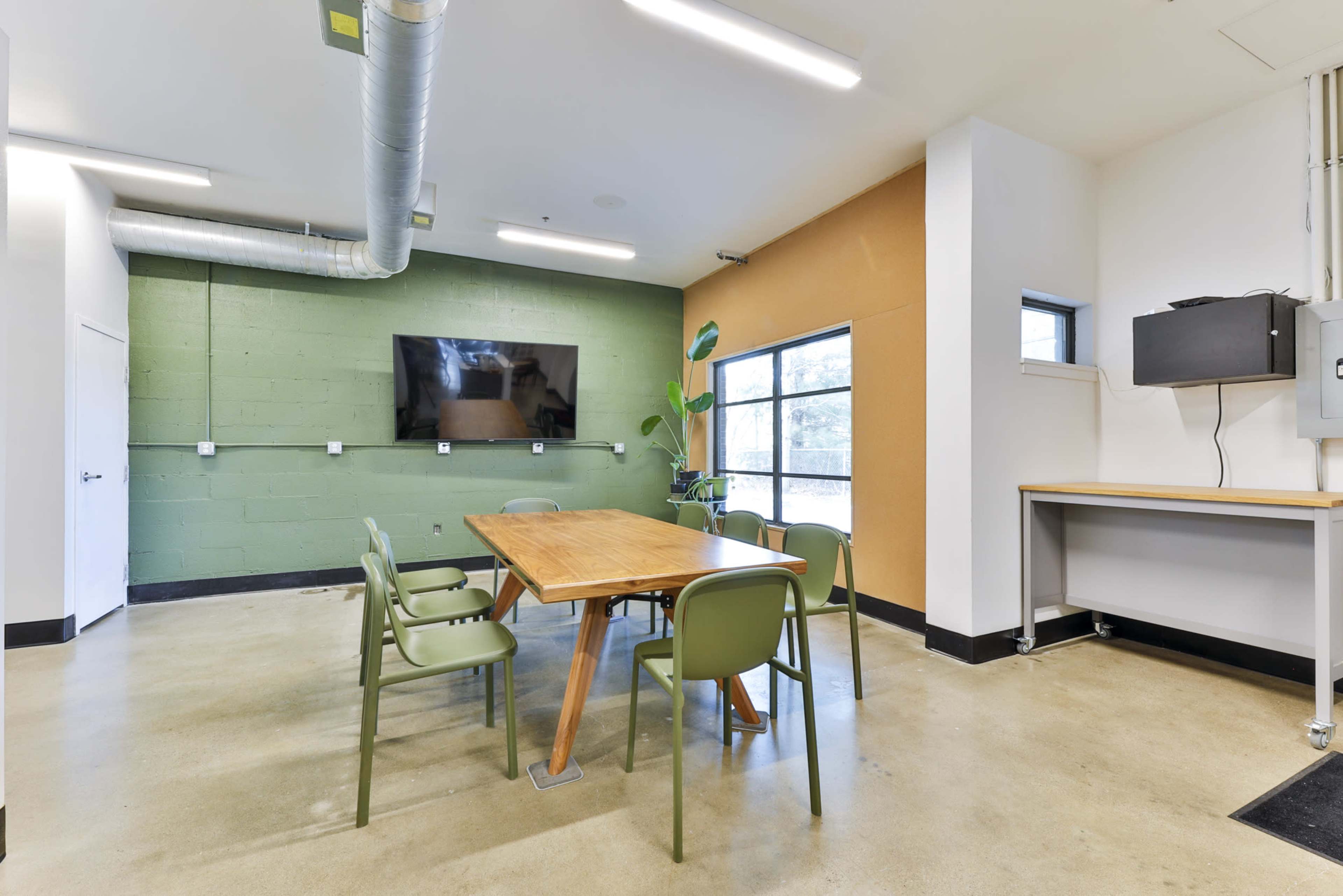 A modern conference room features a wooden table surrounded by green chairs, a wall-mounted television, and large windows.