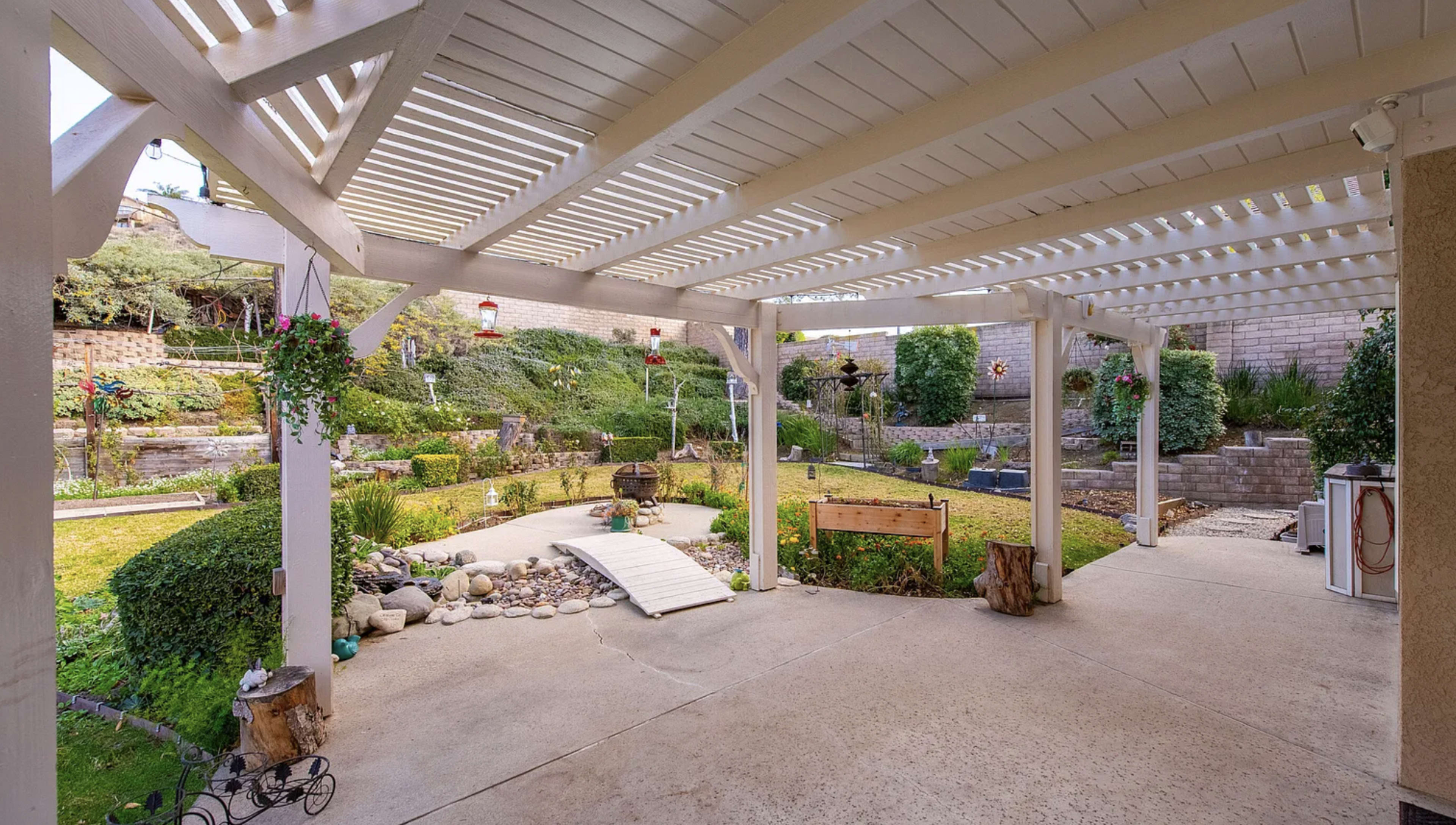 The image shows a spacious outdoor patio with a pergola, surrounded by a well-maintained garden featuring greenery, rocks, and a pathway.