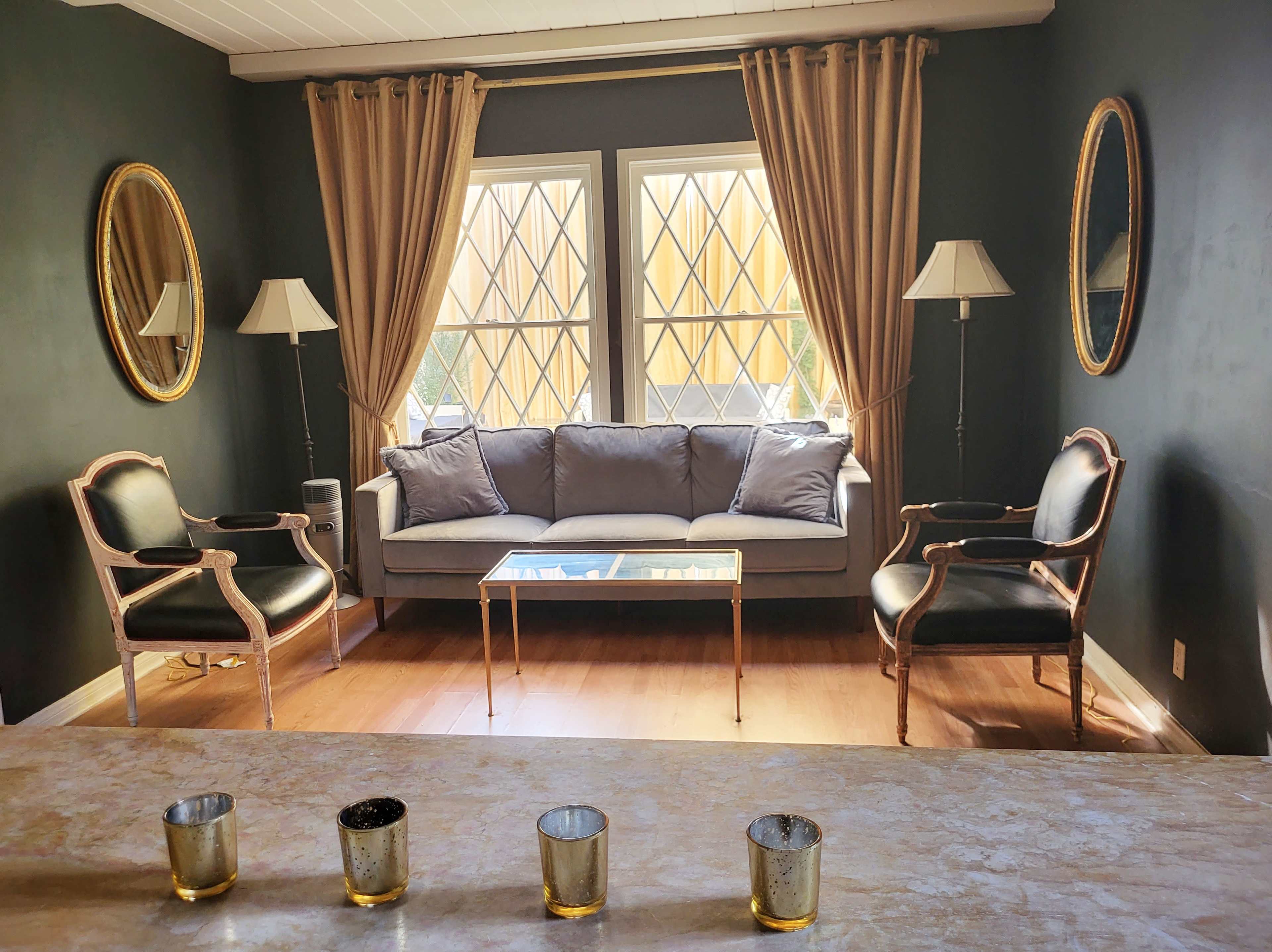 The image shows a living room with a gray sofa, two black armchairs, a glass coffee table, and large windows framed by brown curtains.
