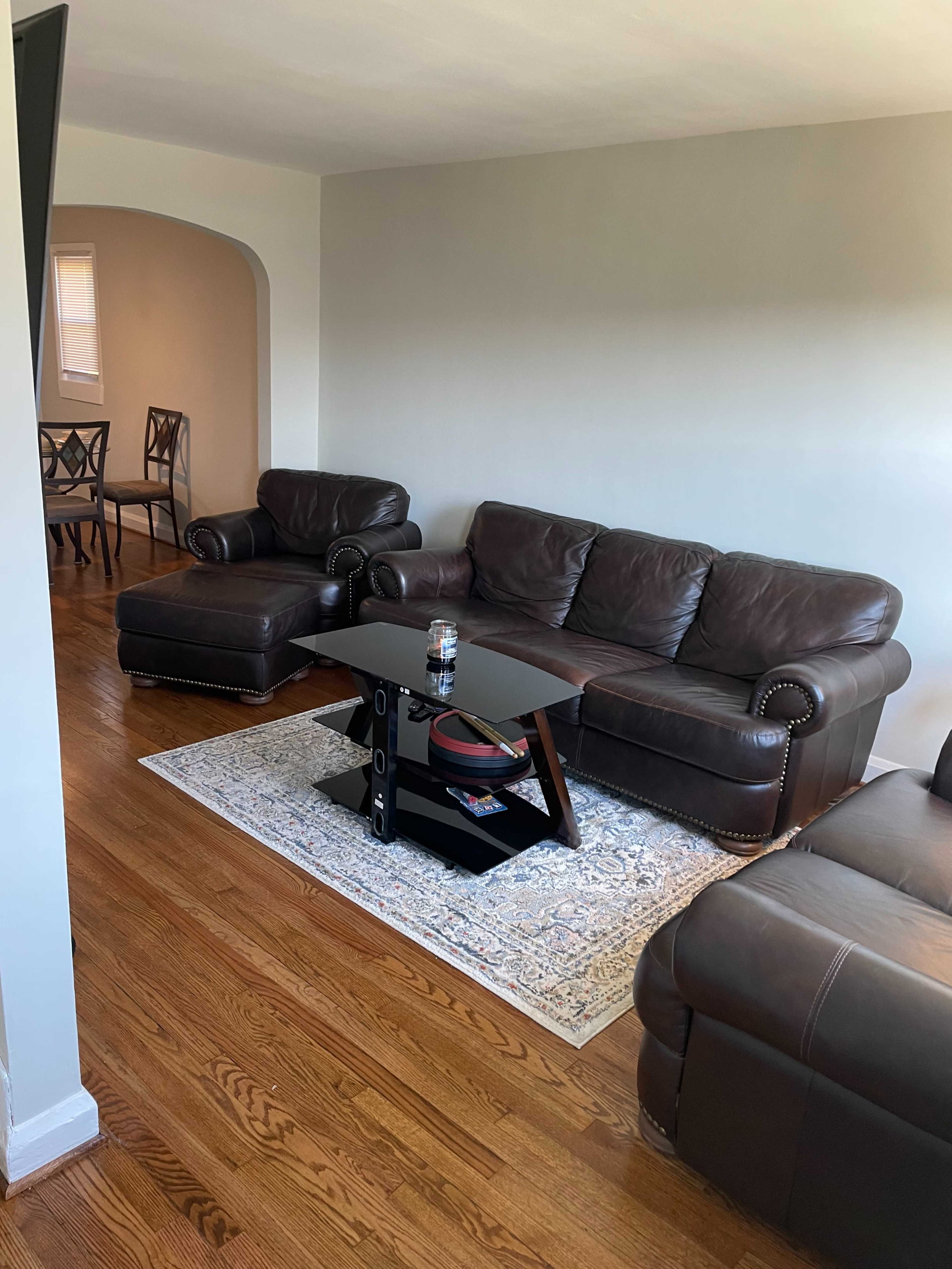 The living room features a brown leather sectional sofa, a glass-top coffee table, and a light-colored area rug on hardwood flooring.