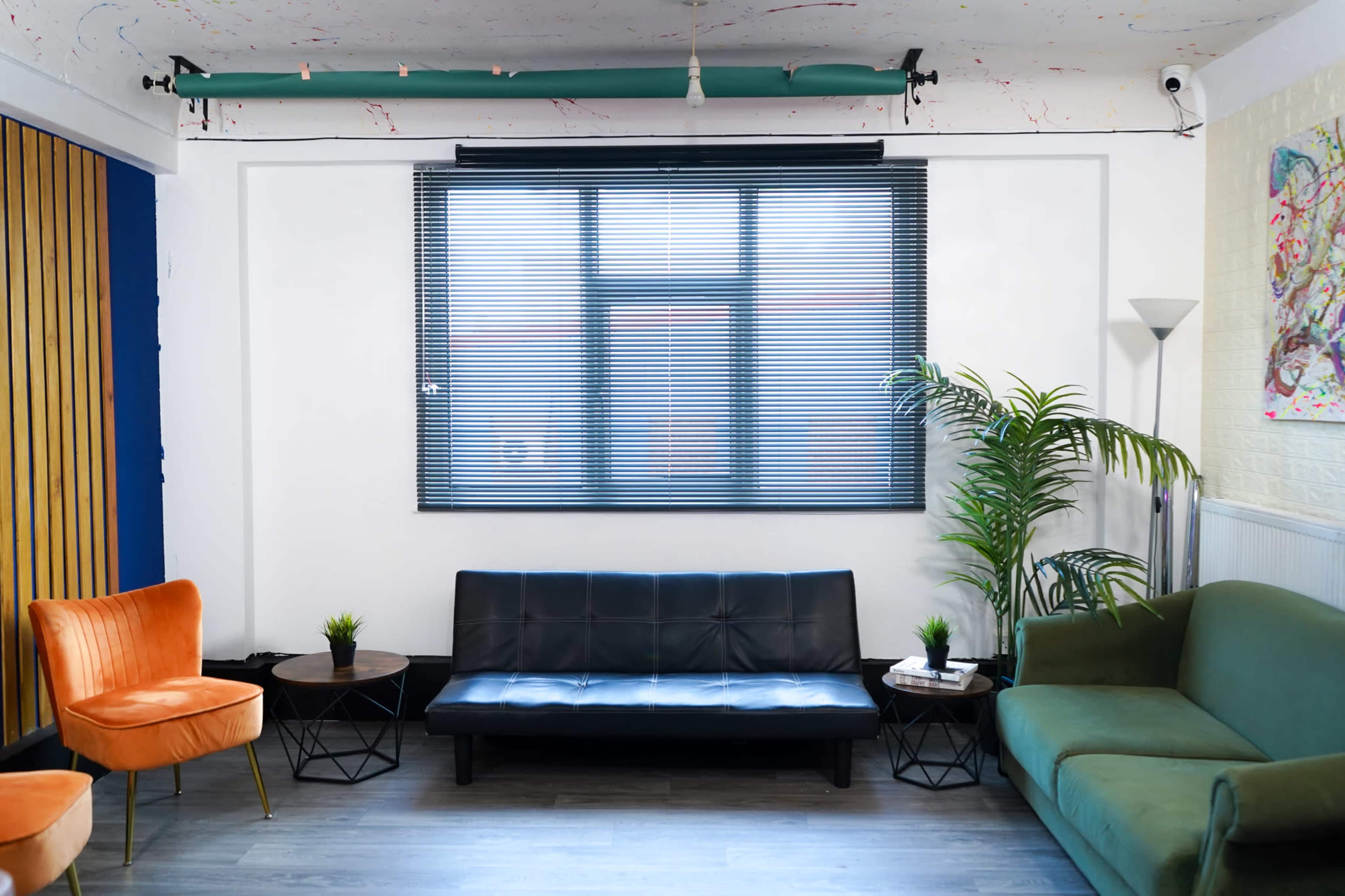 A lounge area with a black sofa, two small side tables, green and orange chairs, and a large window with blinds.