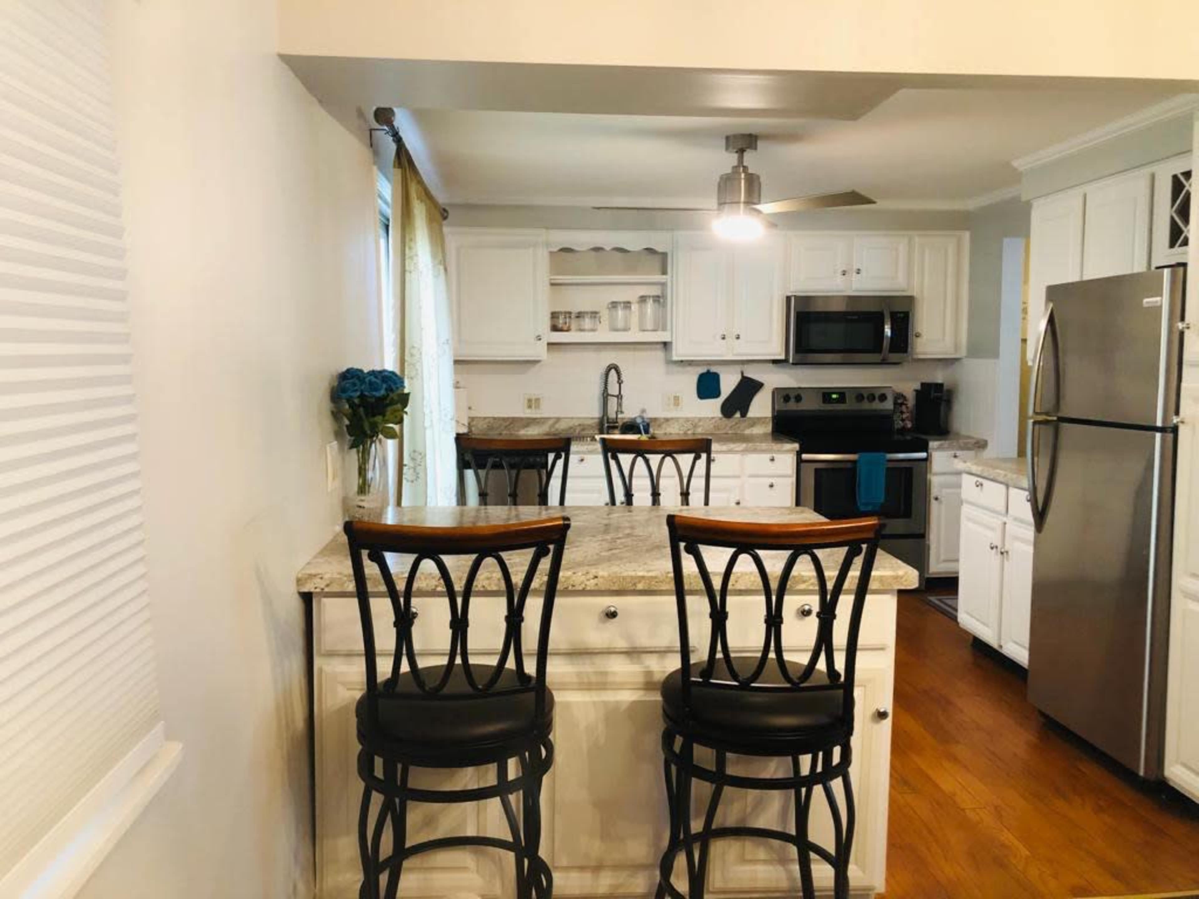 A bright kitchen features white cabinetry, granite countertops with two black bar stools, and modern appliances.
