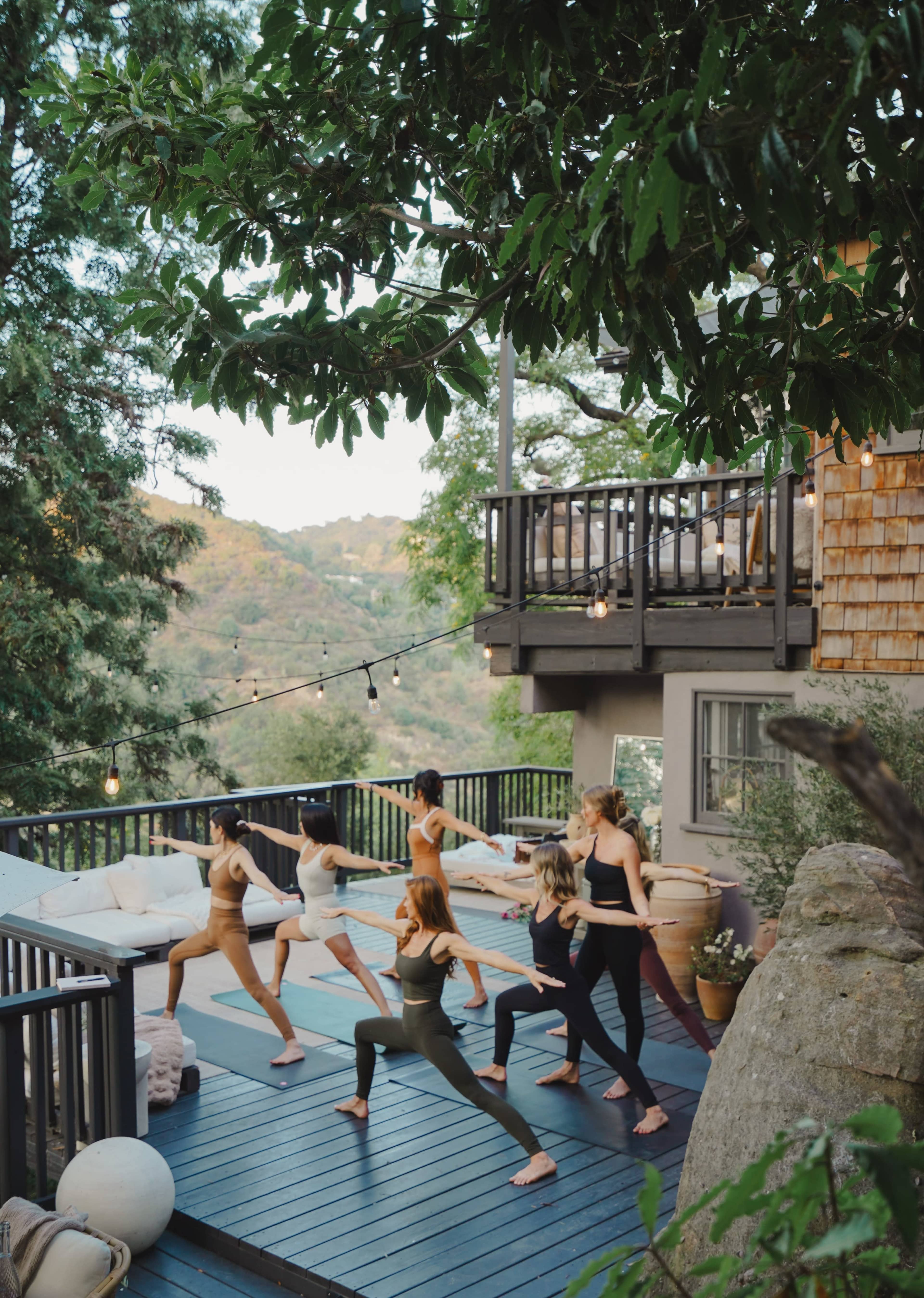 A group of six women perform yoga poses on a deck surrounded by trees, with a scenic view in the background.