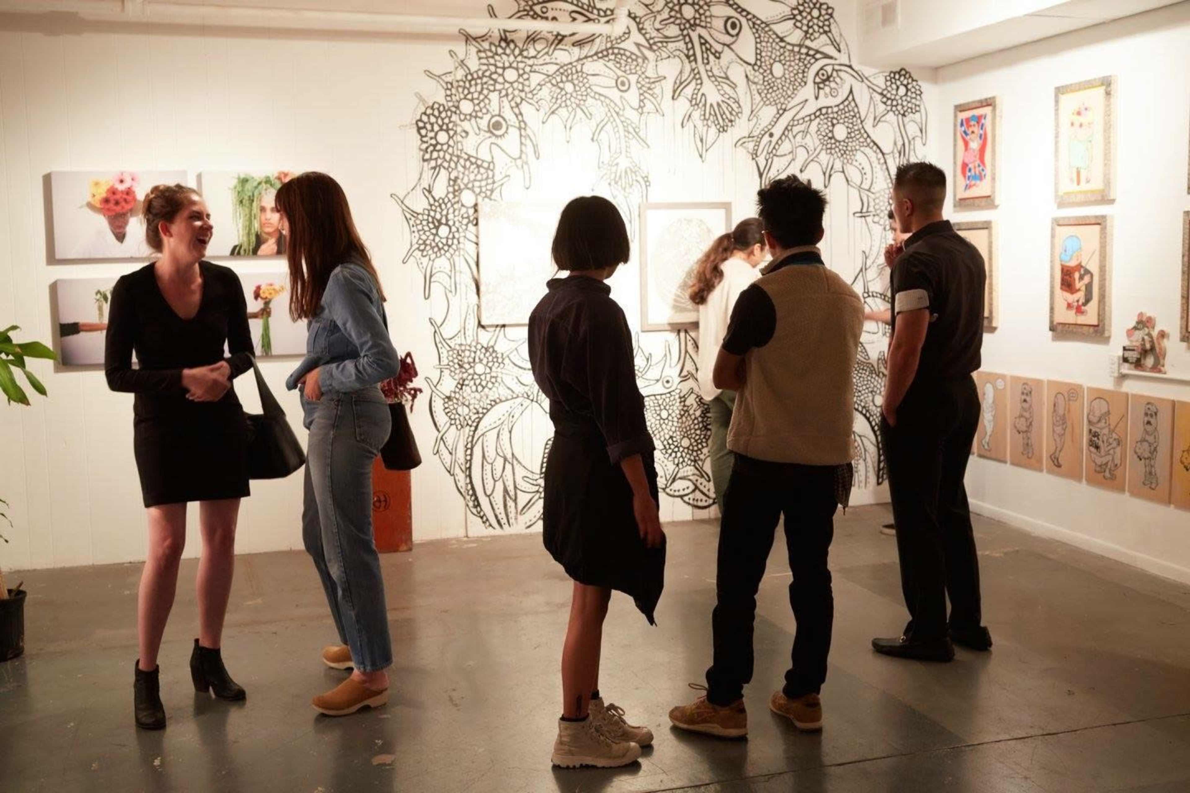 A group of people examines artwork in an art gallery, surrounded by various framed pieces and a mural on the wall.
