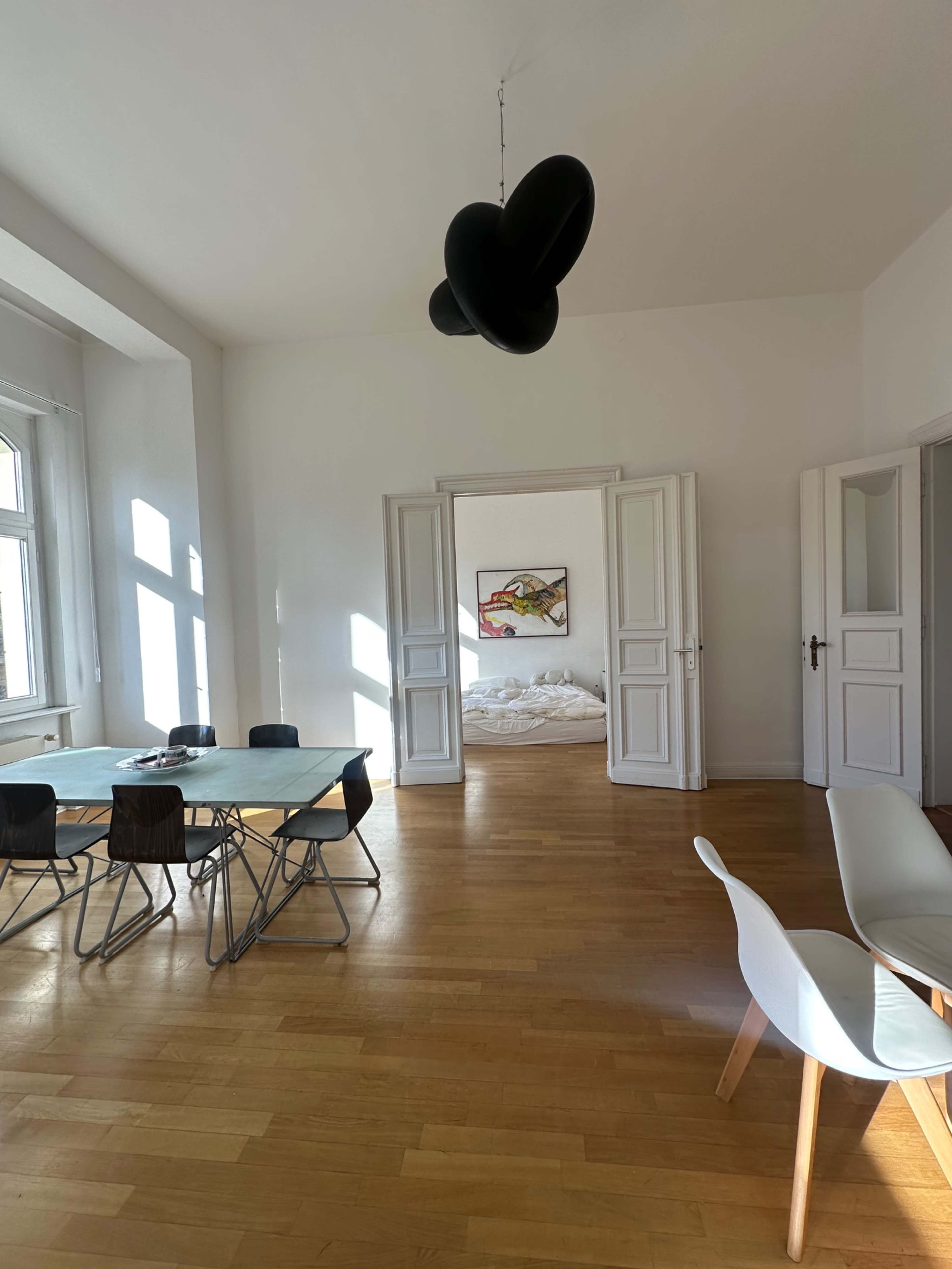 A bright, minimalistic room features a dining table with chairs on one side and a bed with unmade sheets visible through an open doorway.