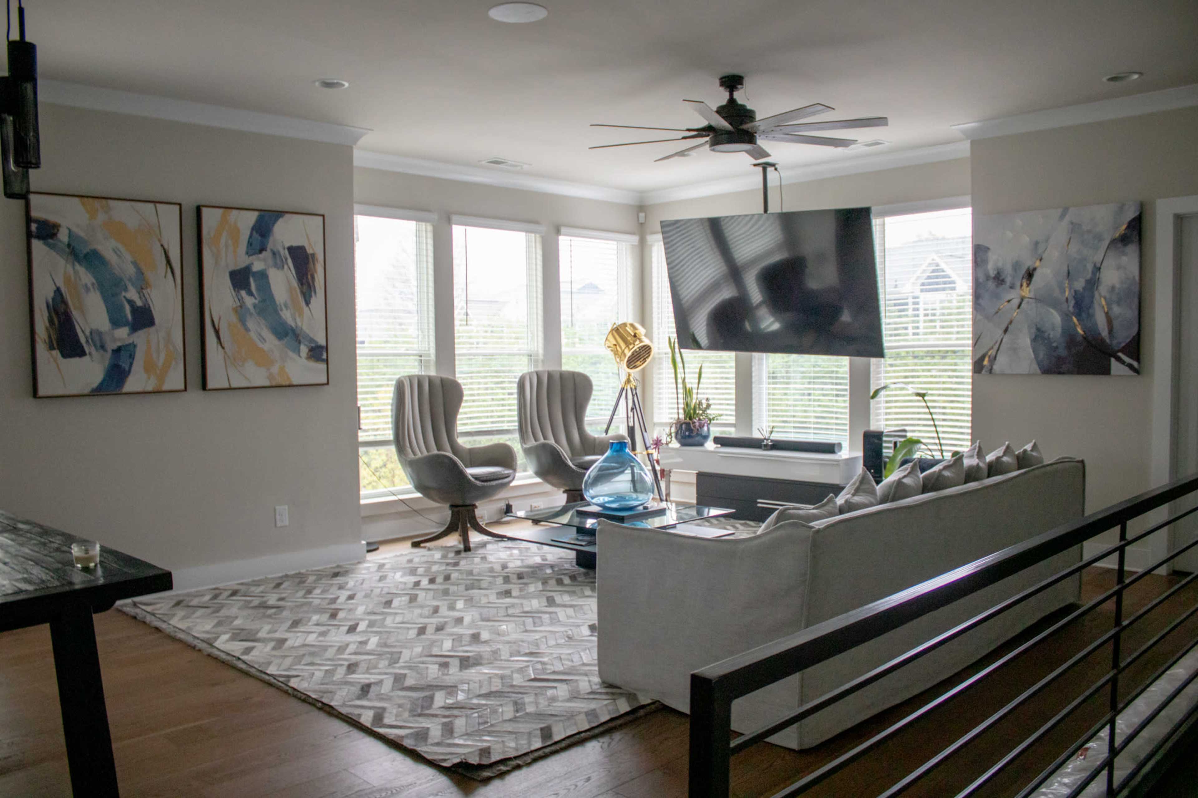A modern living room with large windows, a stylish sectional sofa, two armchairs, abstract artwork on the walls, and a large flat-screen TV mounted above a console table.
