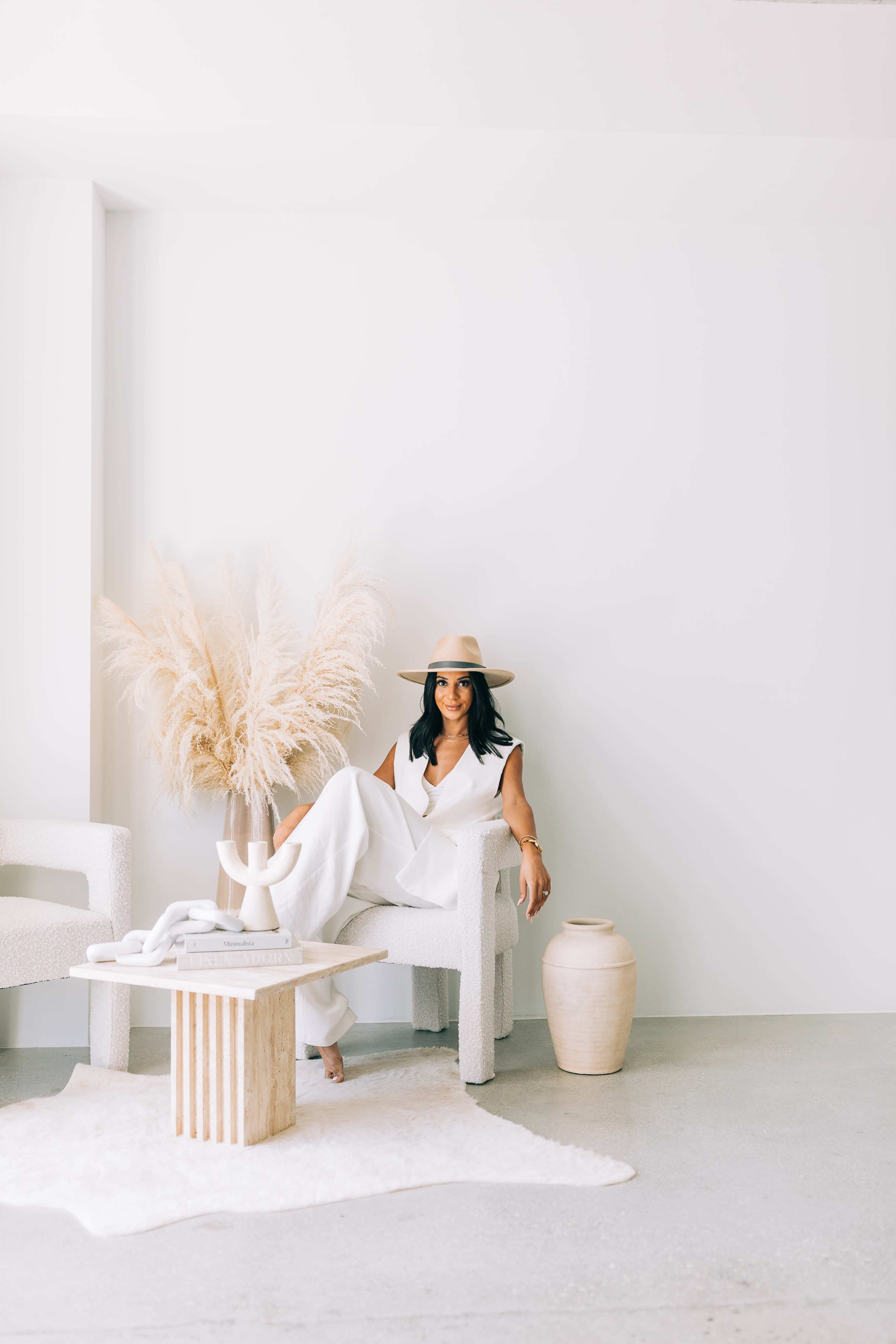 A woman in a white outfit sits on a textured chair beside a large vase and a decorative arrangement of dried plants against a minimalist backdrop.