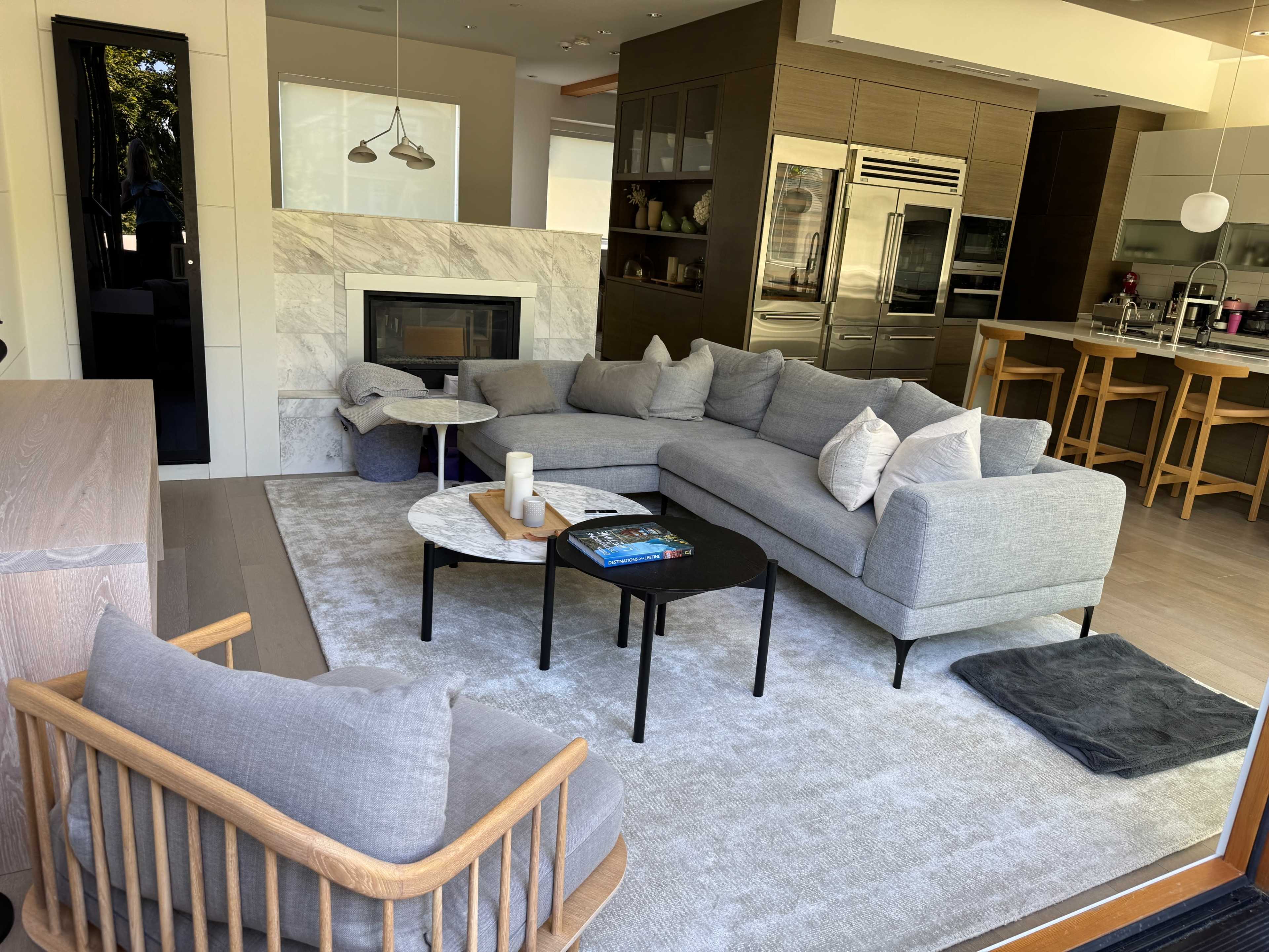 The image shows a modern living room with a large gray sectional sofa, a coffee table, and a view of an open kitchen with stainless steel appliances.