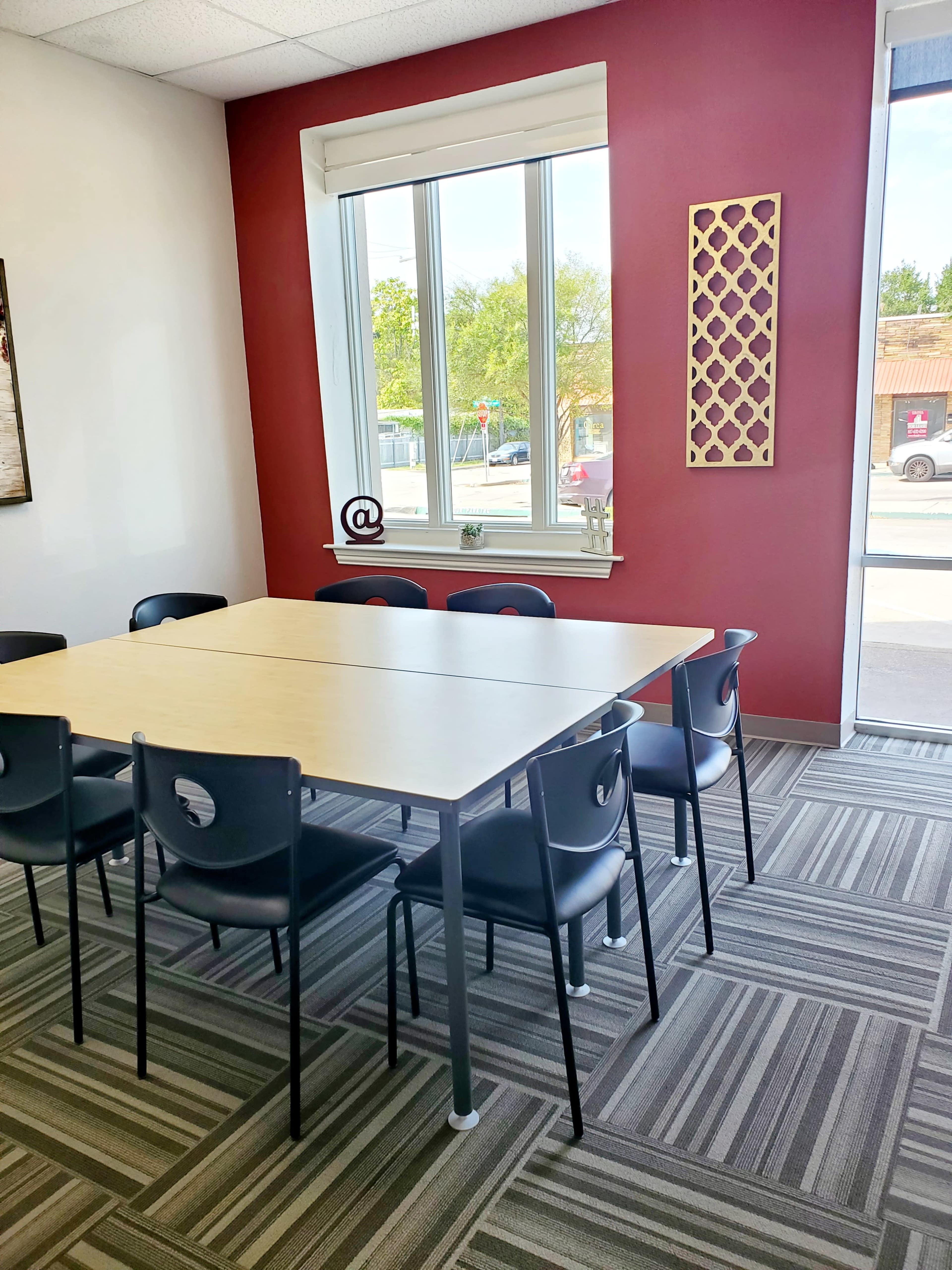 A meeting room features a large rectangular table surrounded by six black chairs against a red accent wall, with large windows letting in natural light.