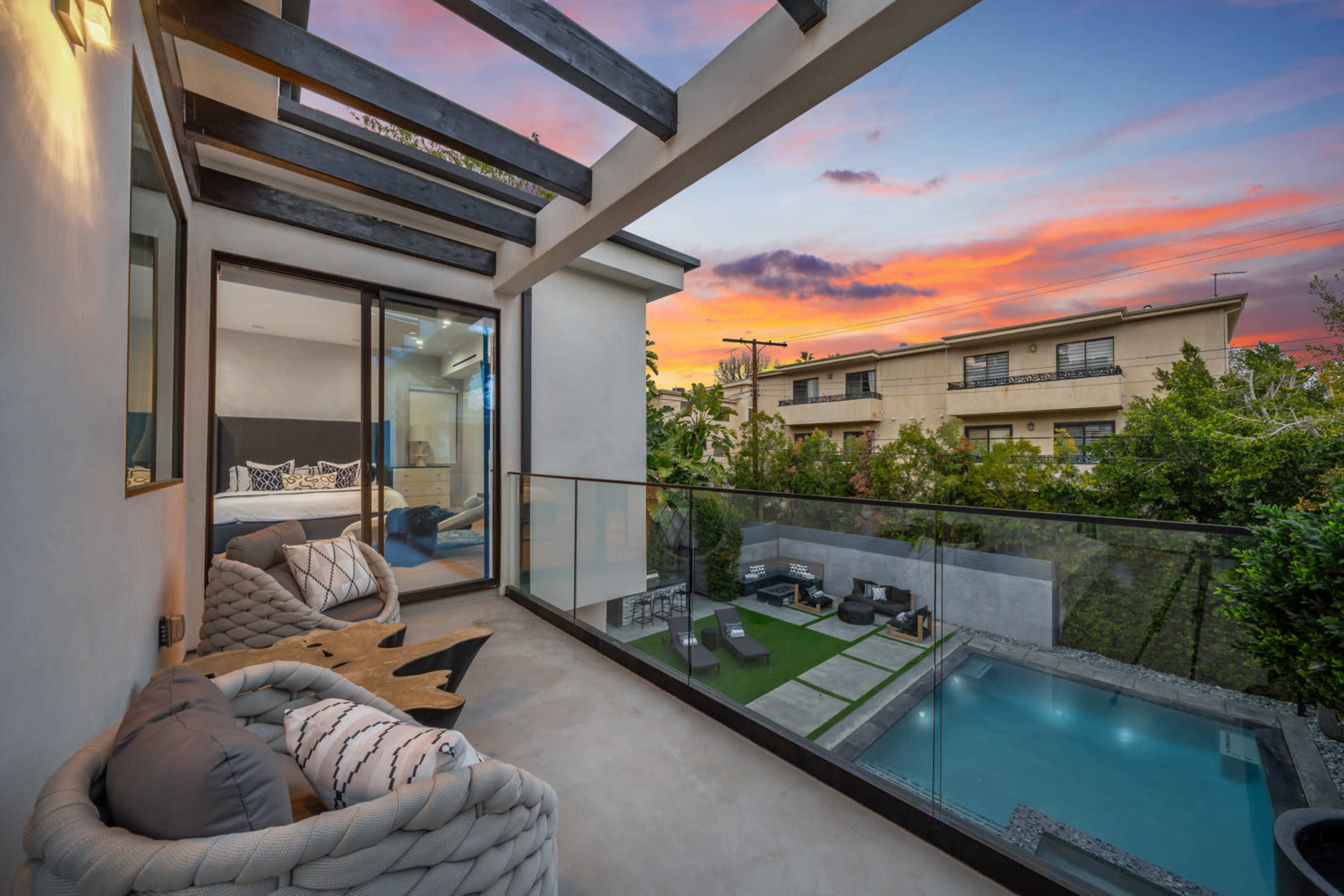 A balcony with modern seating overlooks a backyard pool and landscaped yard against a colorful sunset sky.