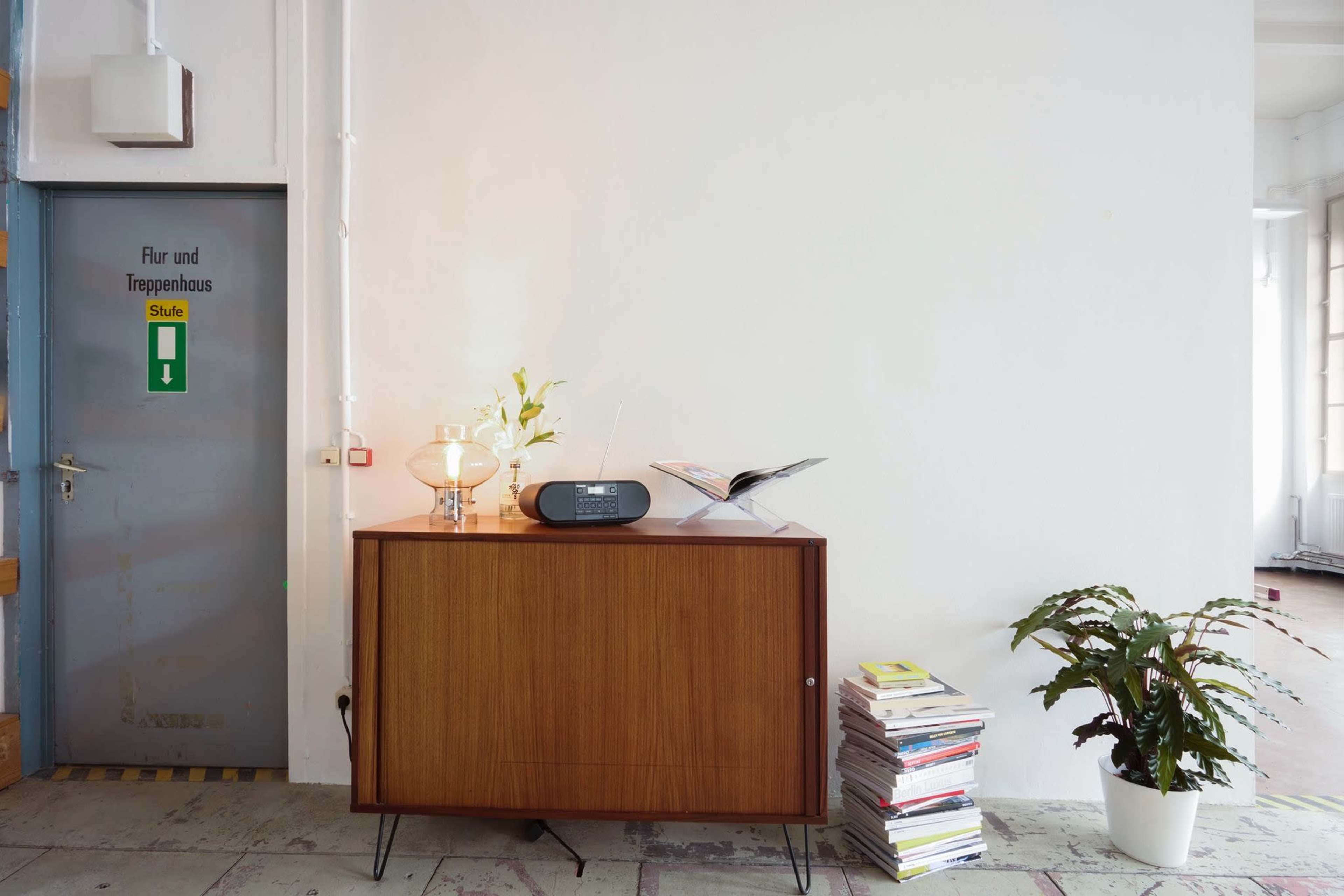 The image shows a minimalist interior with a wooden cabinet, a radio, a small lamp, magazines stacked on the floor, and a potted plant near a door labeled "Flur und Treppenhaus."