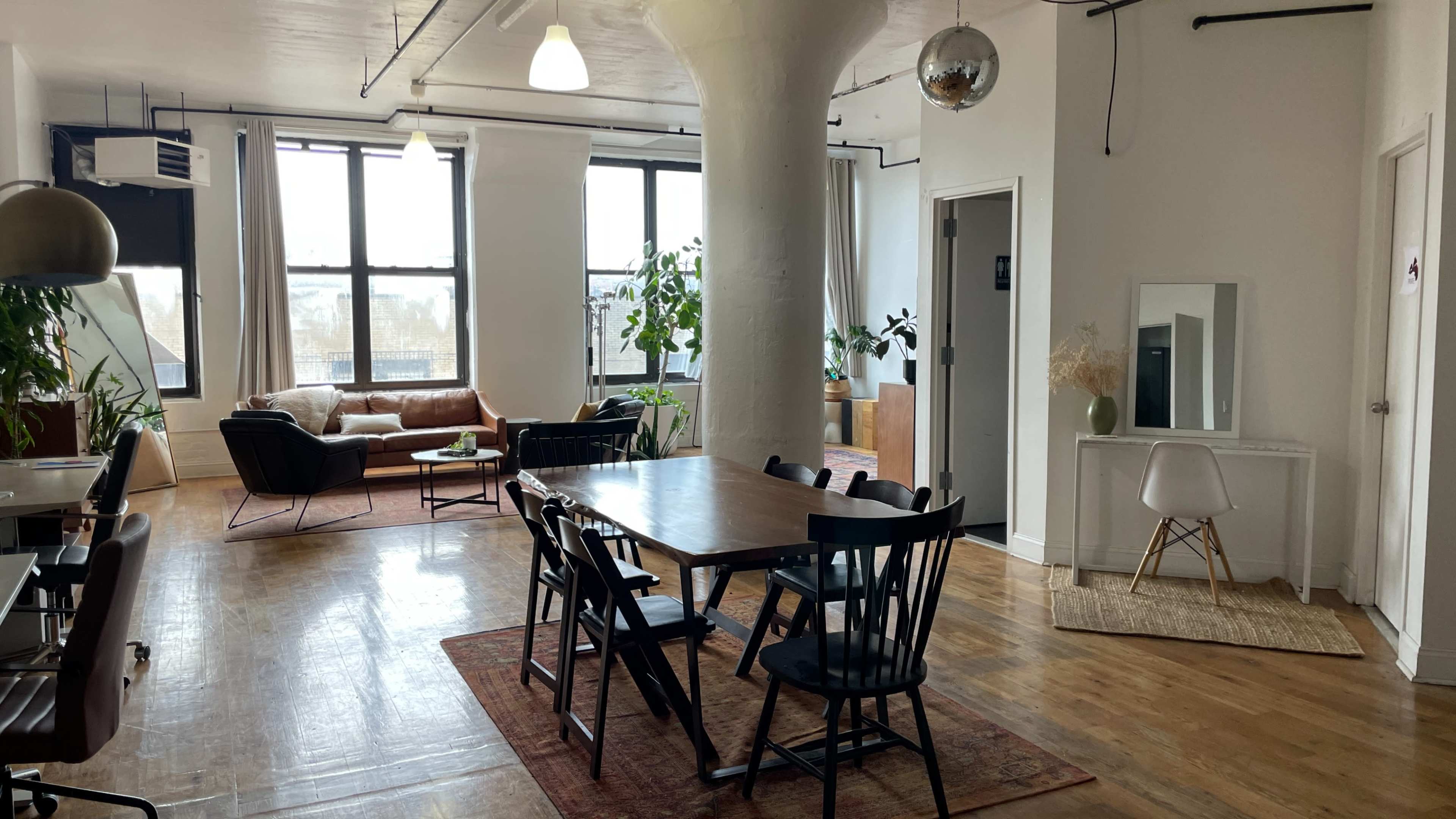 The image shows a spacious, well-lit open area featuring a dining table with chairs, a sofa, plants, and large windows.