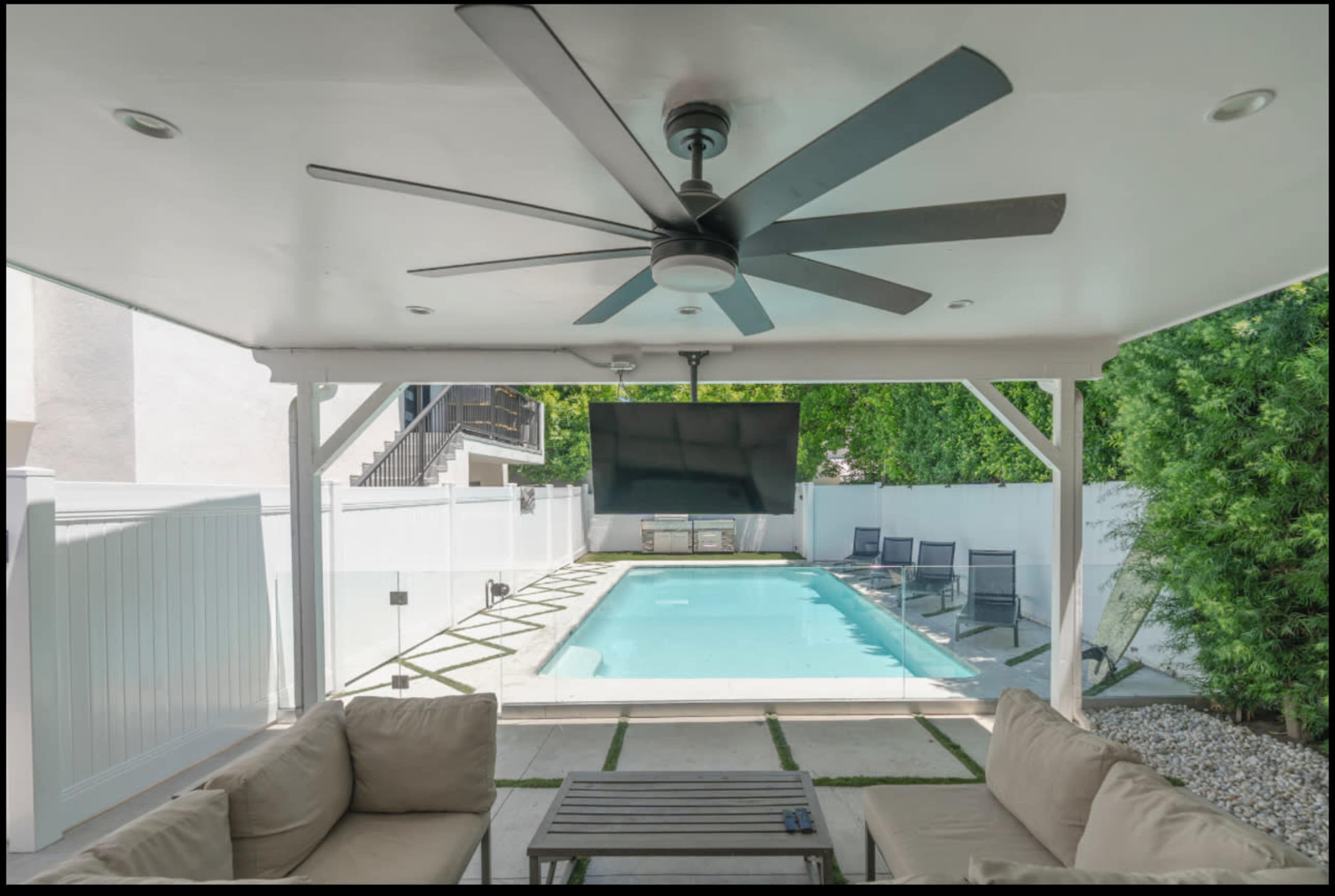 A covered patio features a ceiling fan, a seating area with beige cushions, and a view of a rectangular swimming pool surrounded by a white fence.