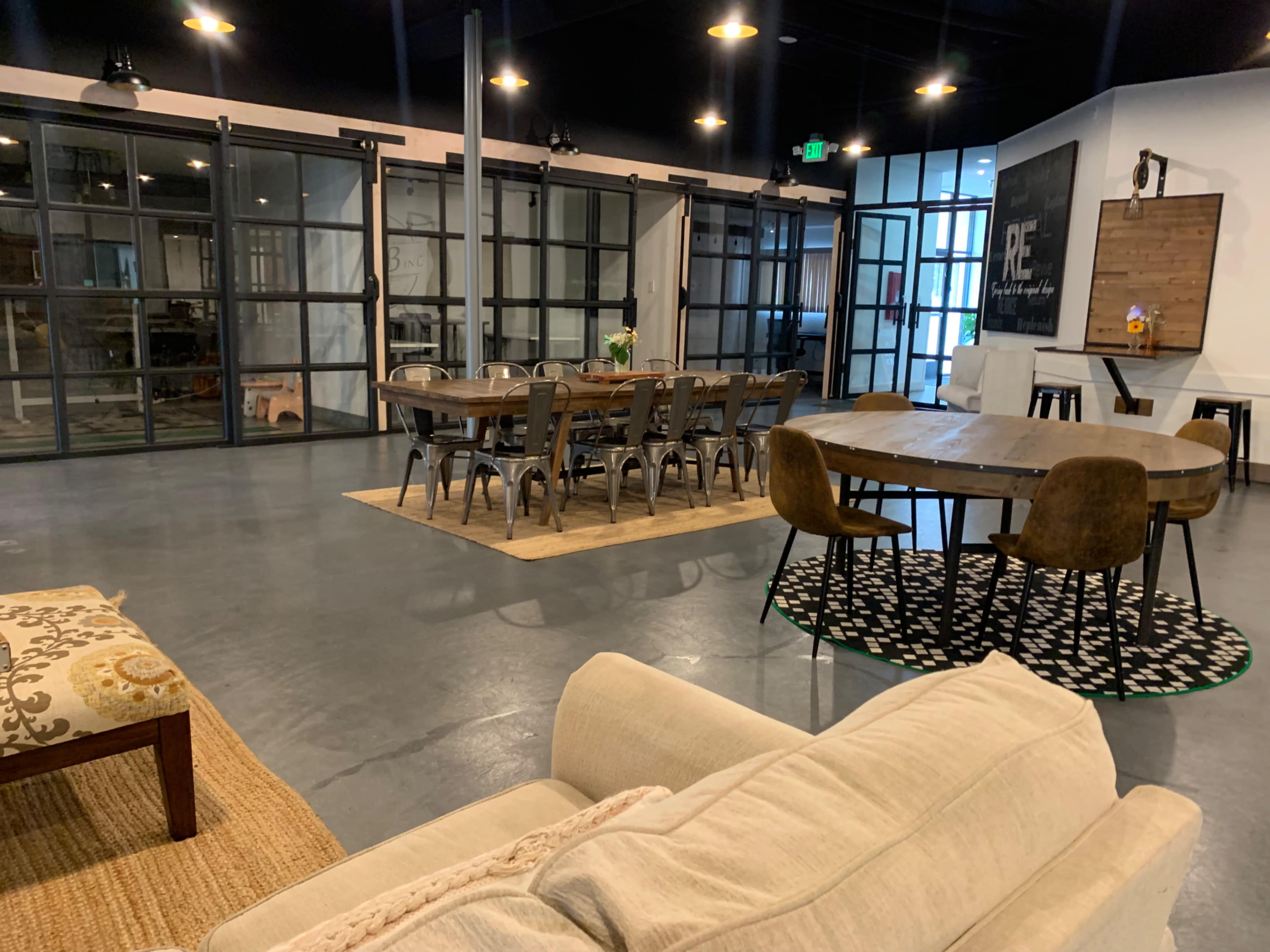 A spacious and modern interior features a mix of seating areas, including a couch, dining tables, and a rug, with large glass partitions separating different zones.