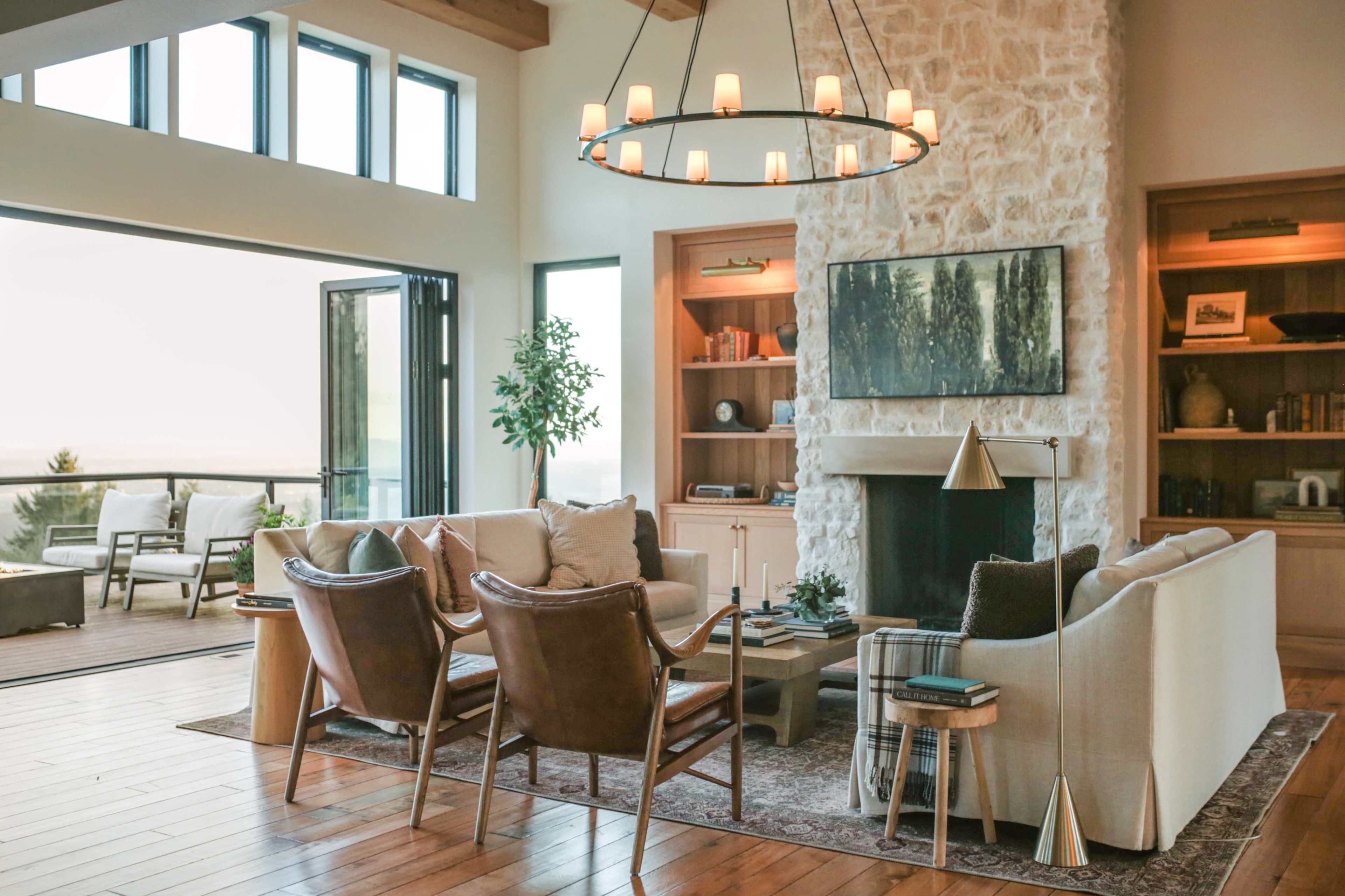 The image shows a modern living room with a large stone fireplace, comfortable seating, and large windows that overlook a scenic view.