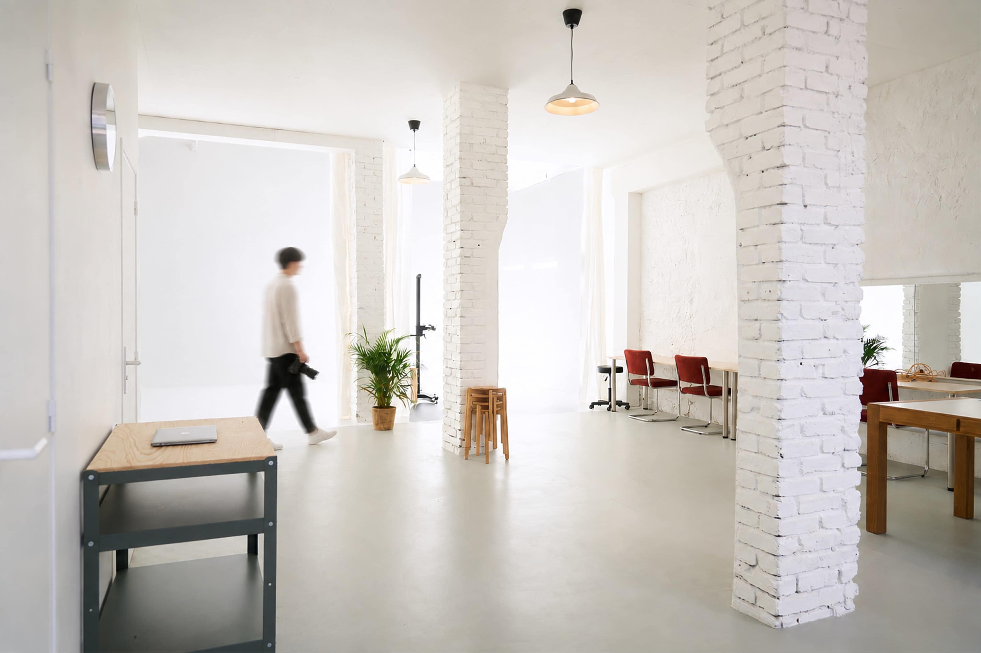 The image shows a modern, bright studio space with white brick pillars, minimal furniture, and a person walking through the area.