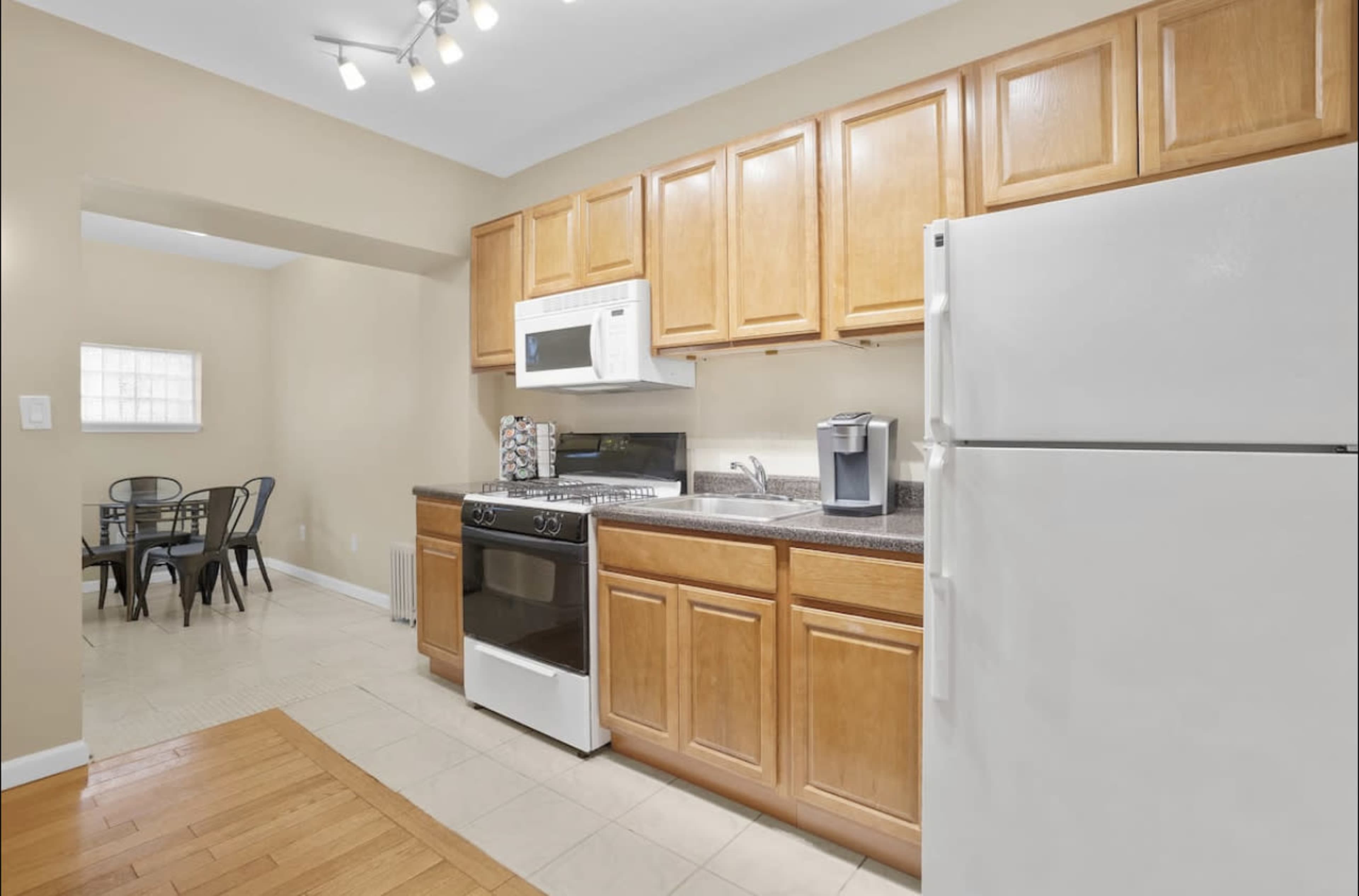 The image shows a kitchen with wooden cabinets, a white refrigerator, a gas stove, a microwave, and a small dining area with a table and chairs in the background.