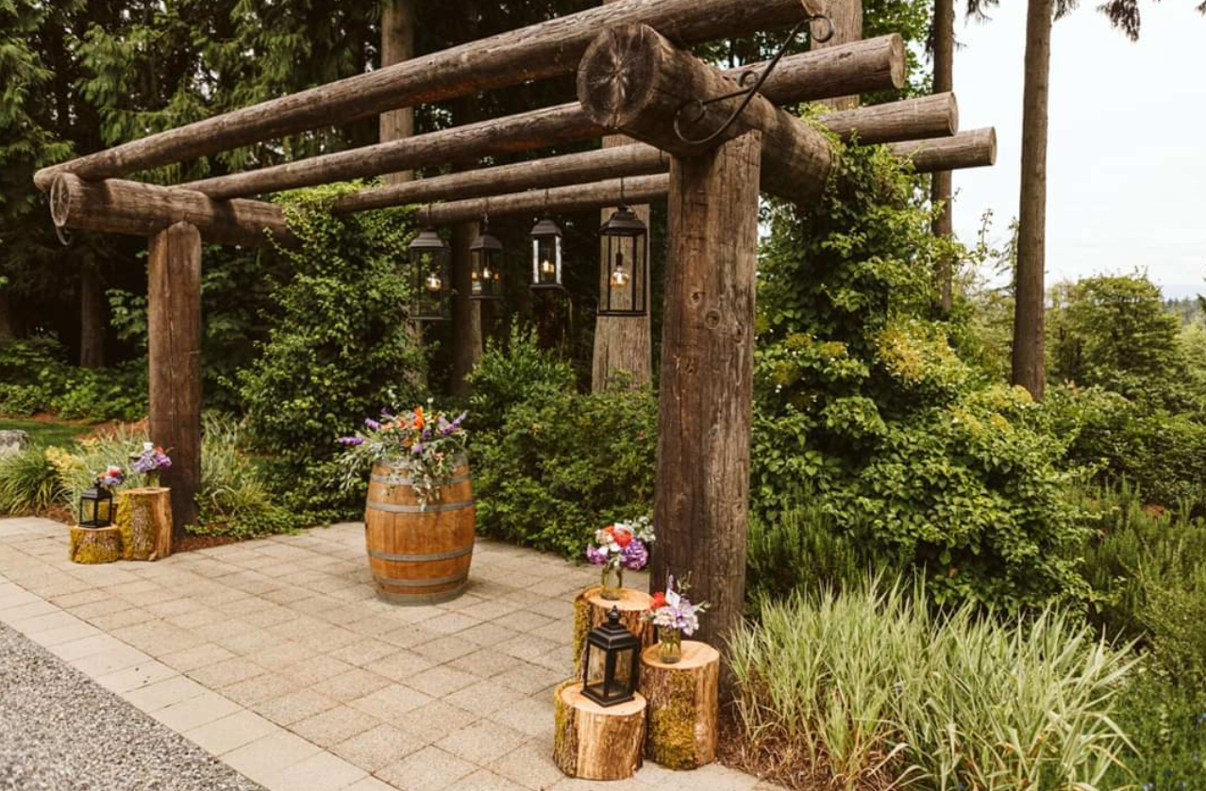 The image shows a rustic wooden archway adorned with lanterns, surrounded by greenery, with a decorative barrel and flower arrangements on either side.