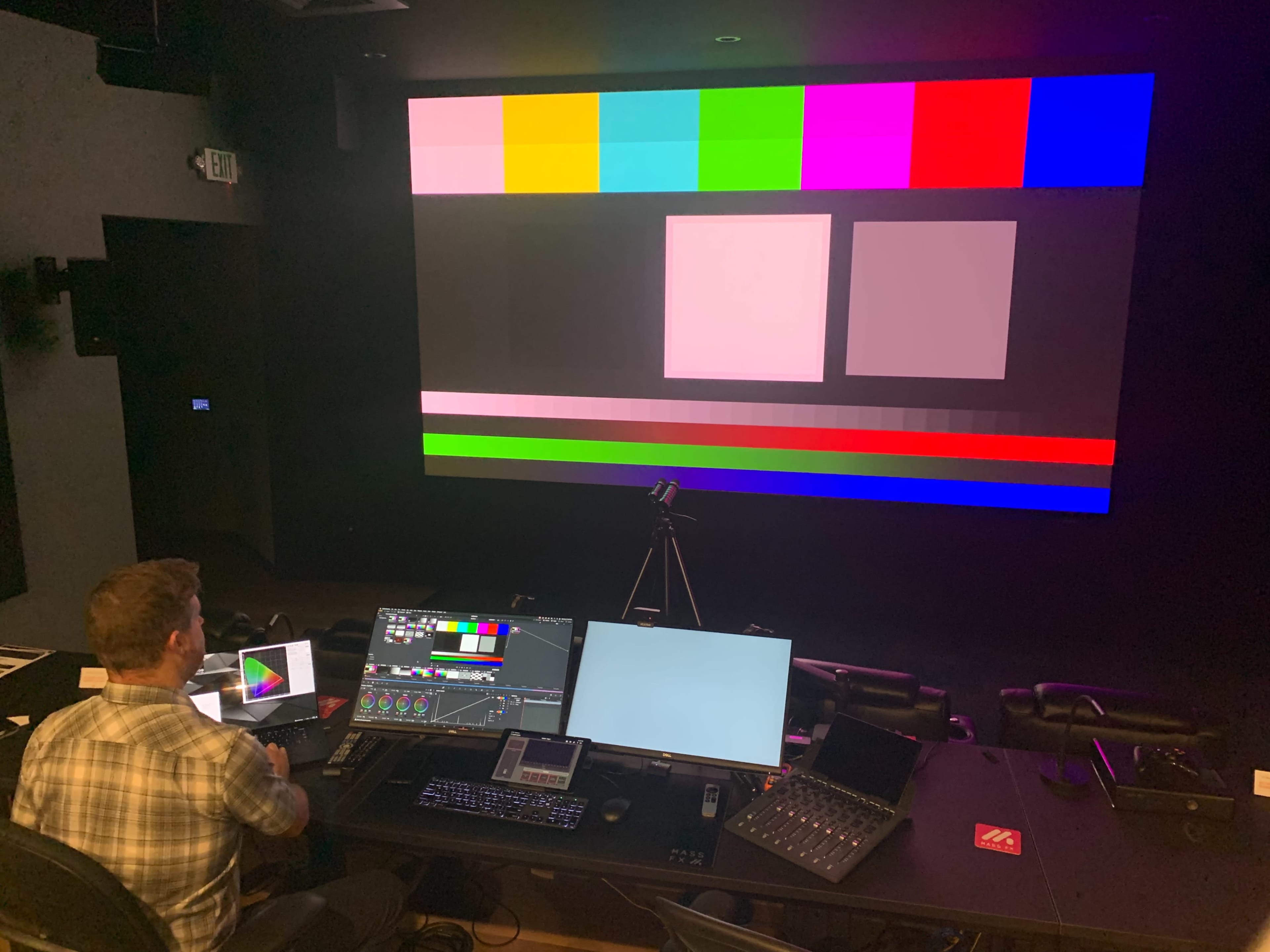 A technician is seated at a control desk in a dimly lit room, adjusting color settings on multiple screens while colorful test patterns are displayed on a large wall monitor.