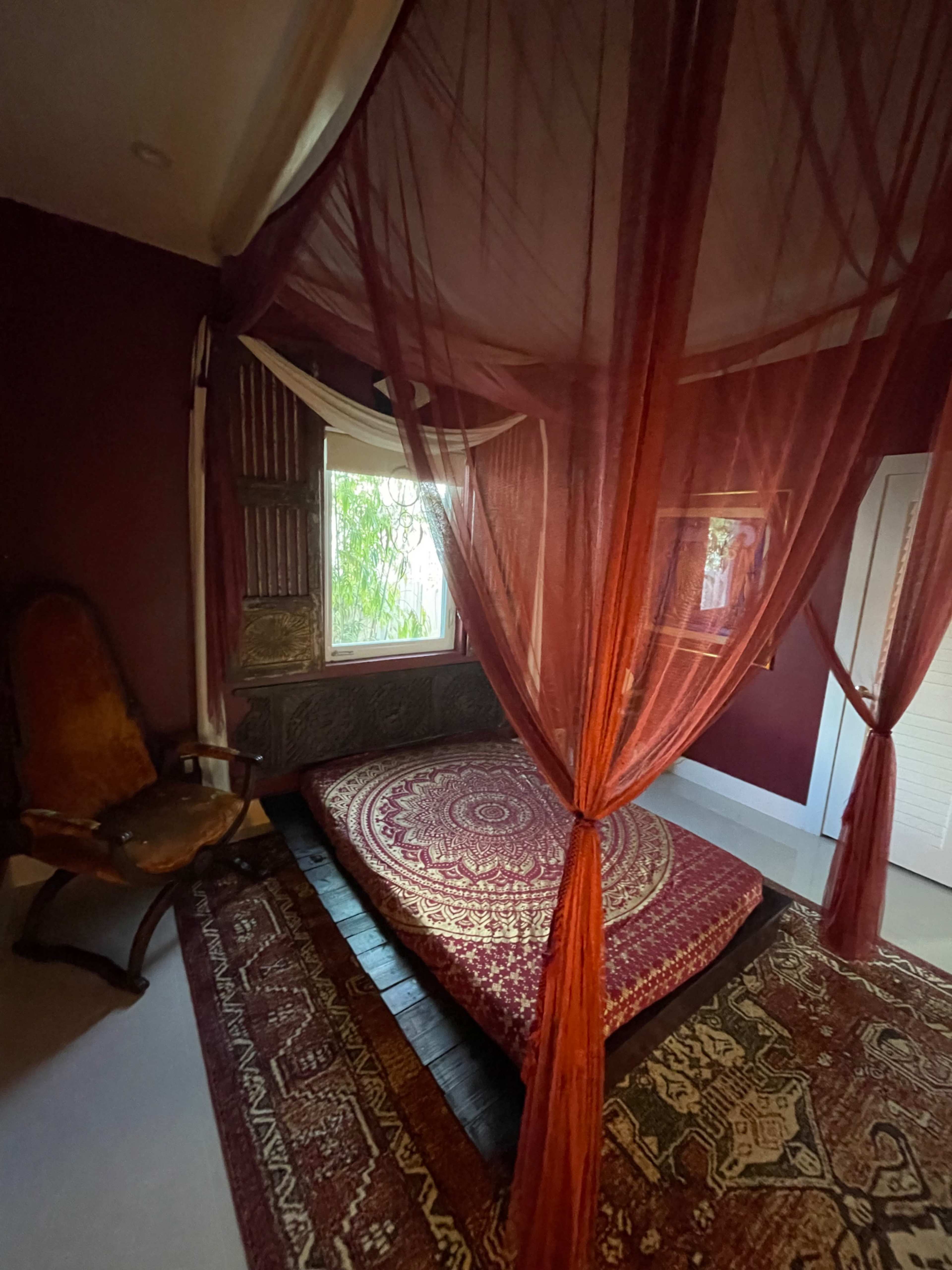 A room with a low, circular bed surrounded by sheer red curtains, a patterned rug on the floor, and a wooden chair in the corner.