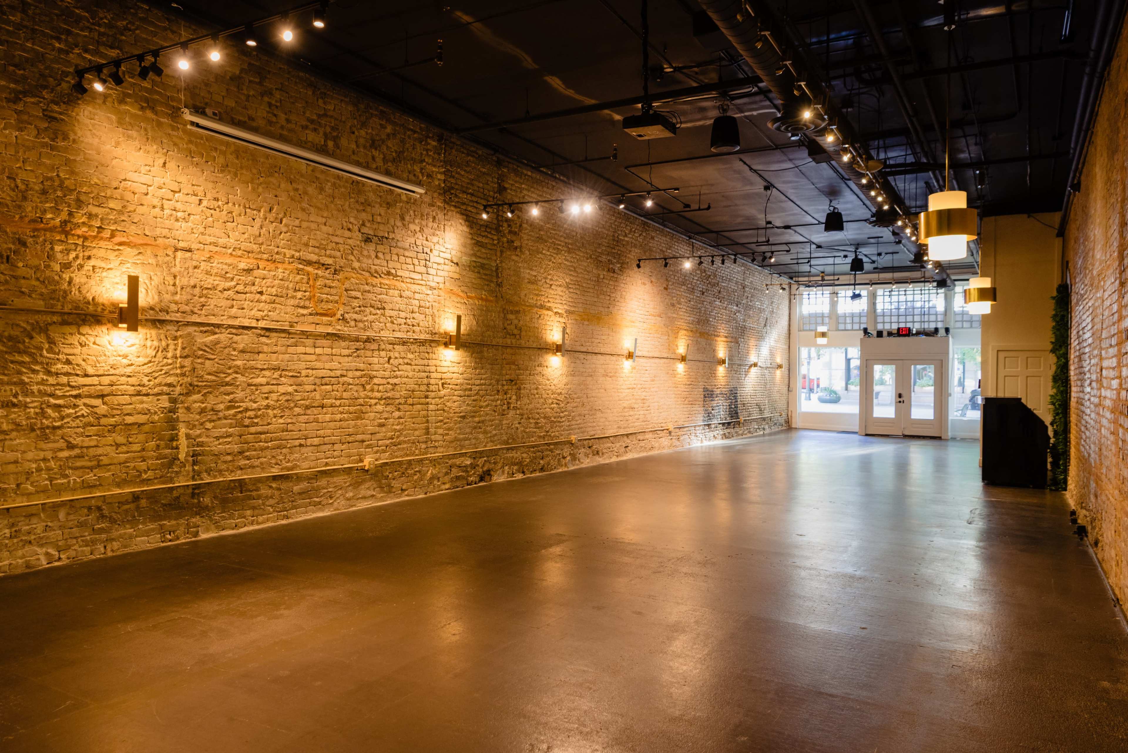 The image shows a spacious, well-lit interior with exposed brick walls and a polished floor, featuring minimal furnishings and a pair of double doors at the far end.
