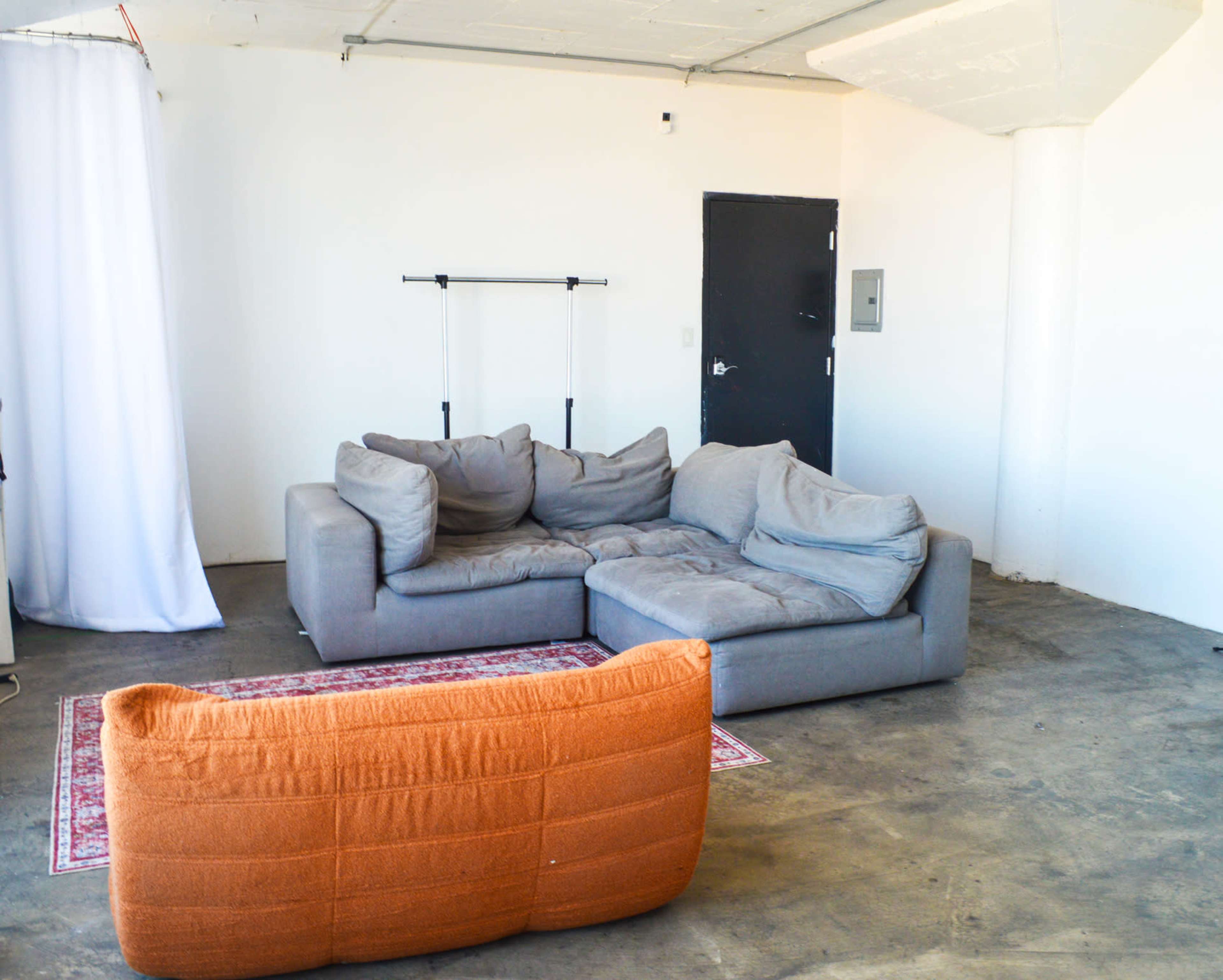 The image shows a spacious, minimalist room with a gray sectional sofa, an orange sofa, and a clothing rack against a white wall.