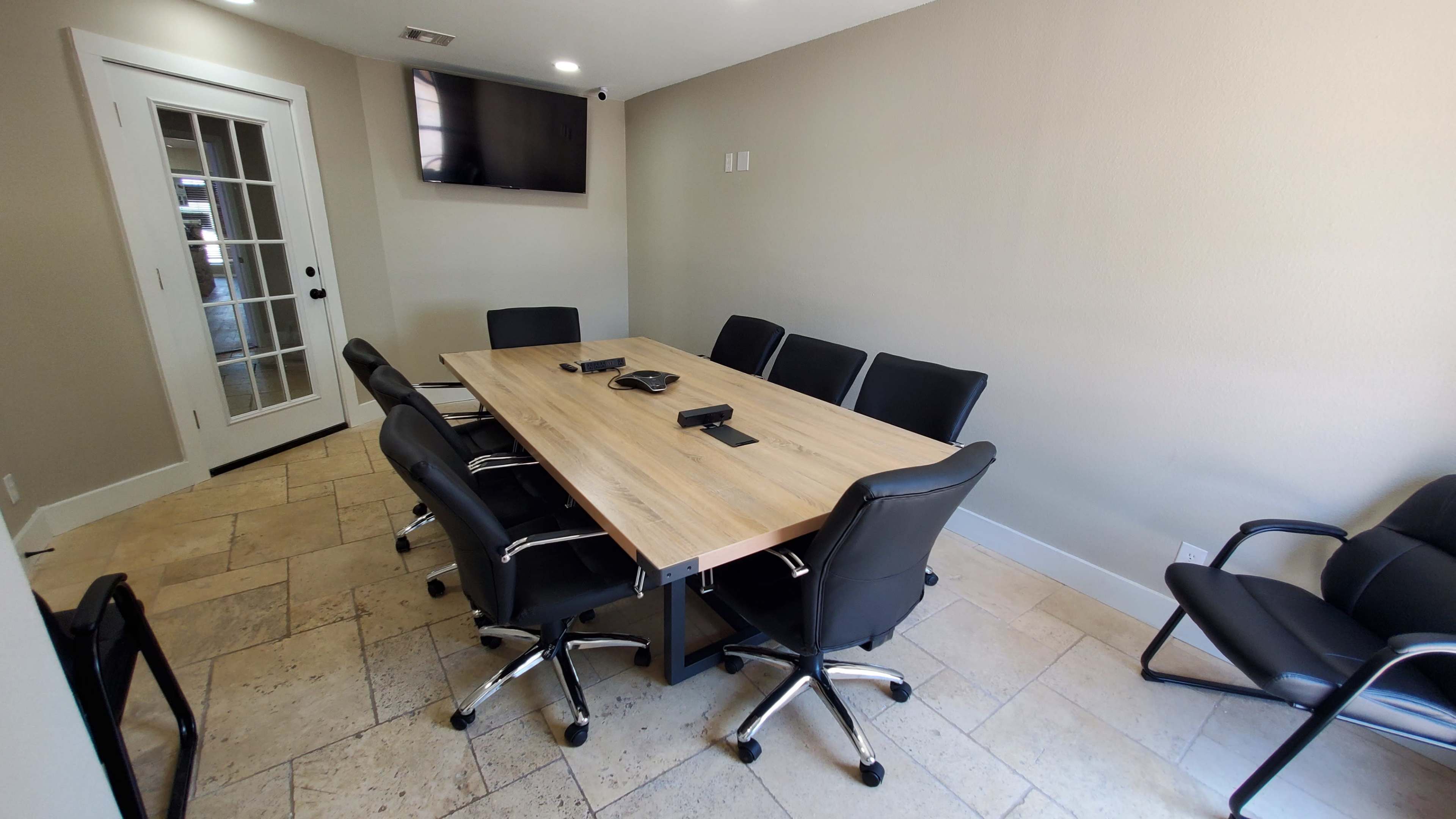 A modern conference room features a large wooden table surrounded by black leather chairs, with a television on the wall and a door leading to an exterior space.