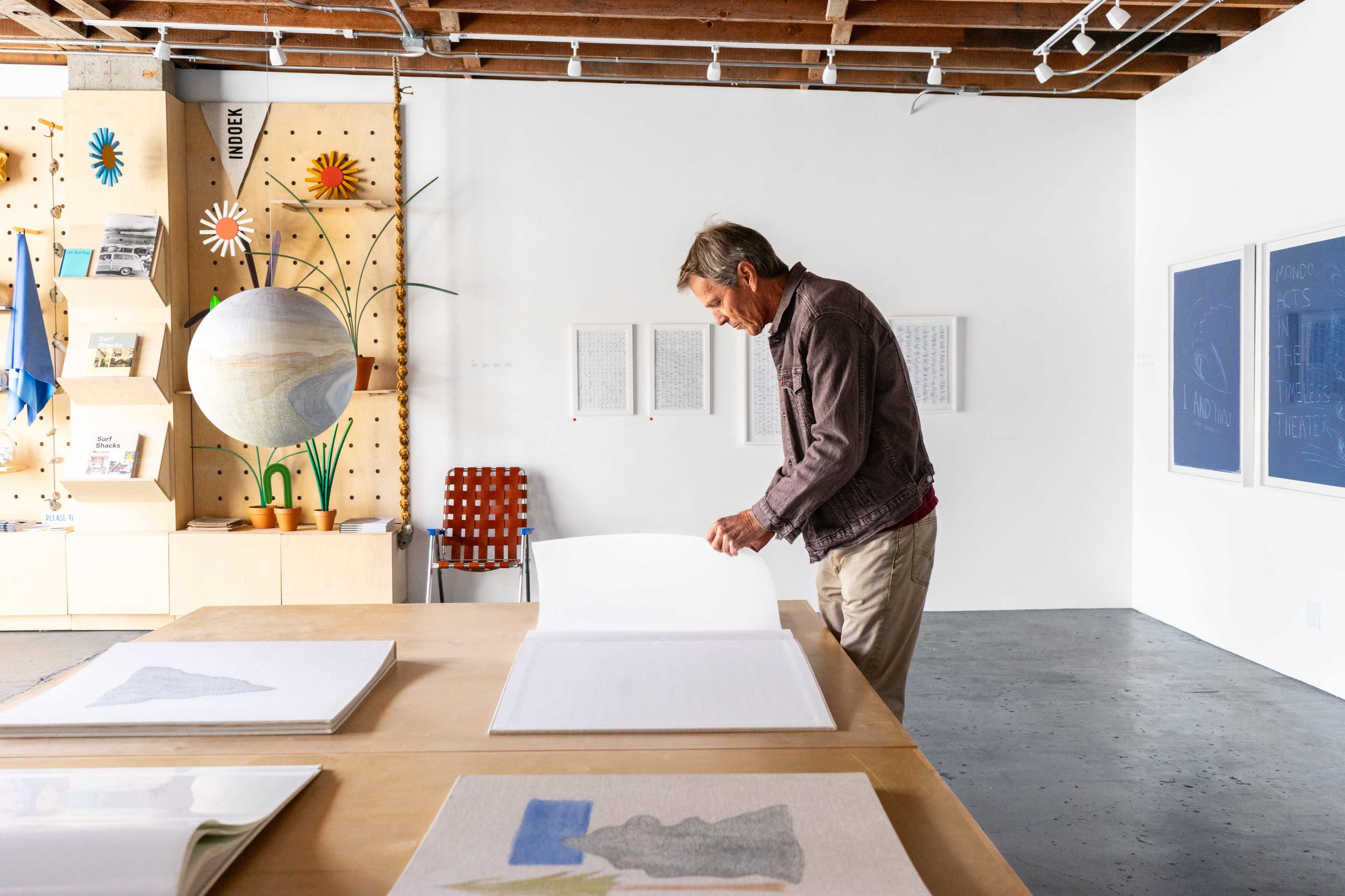 A man stands at a table flipping through large sheets of paper in a brightly lit art studio with various artworks displayed on the walls.