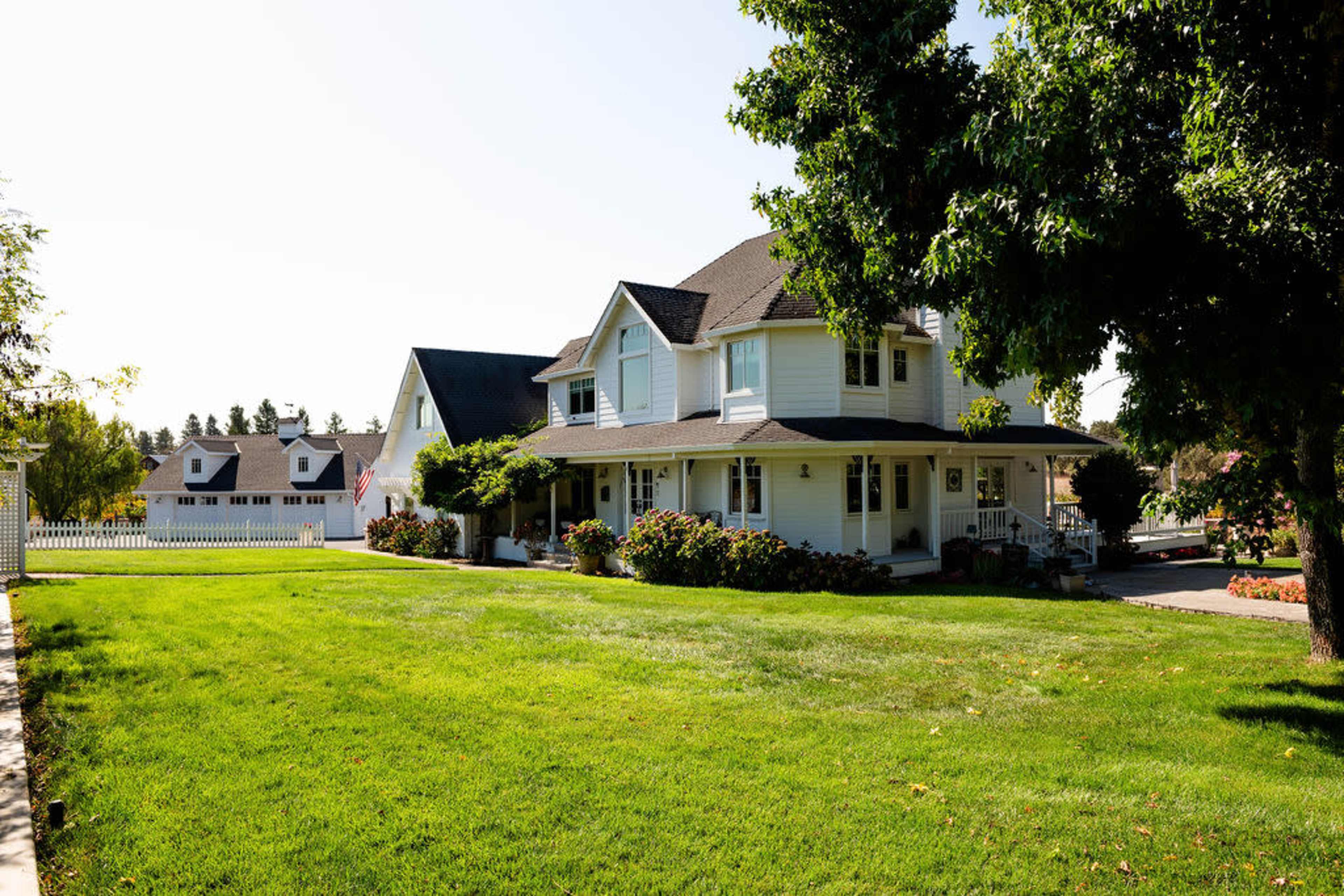 The image shows a two-story white house with a front porch, surrounded by a well-manicured lawn and bushes, with another house visible in the background.