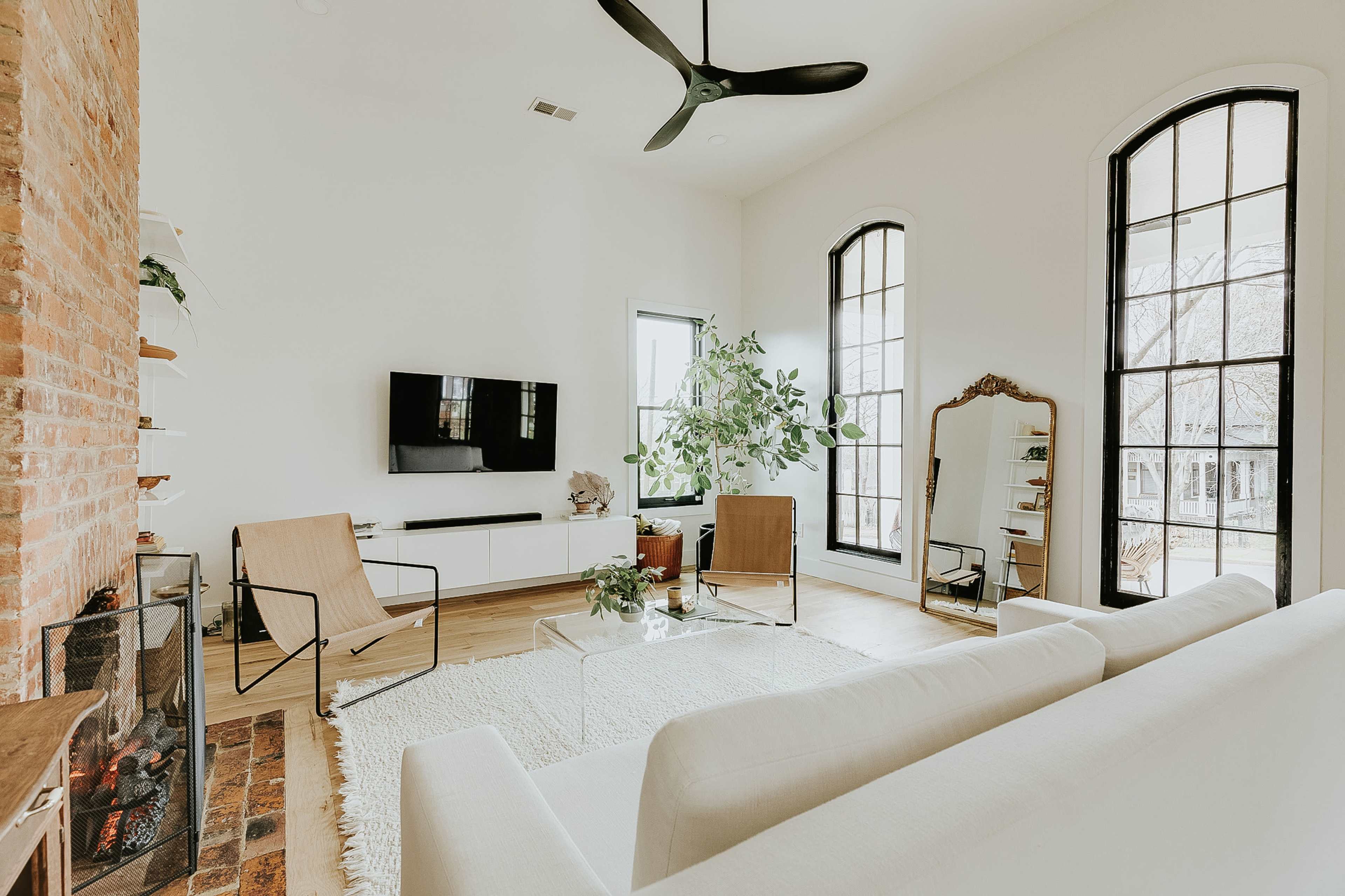 The living room features a white sofa, two chairs, a television mounted on the wall, and large windows, accented by a potted plant and a mirror.