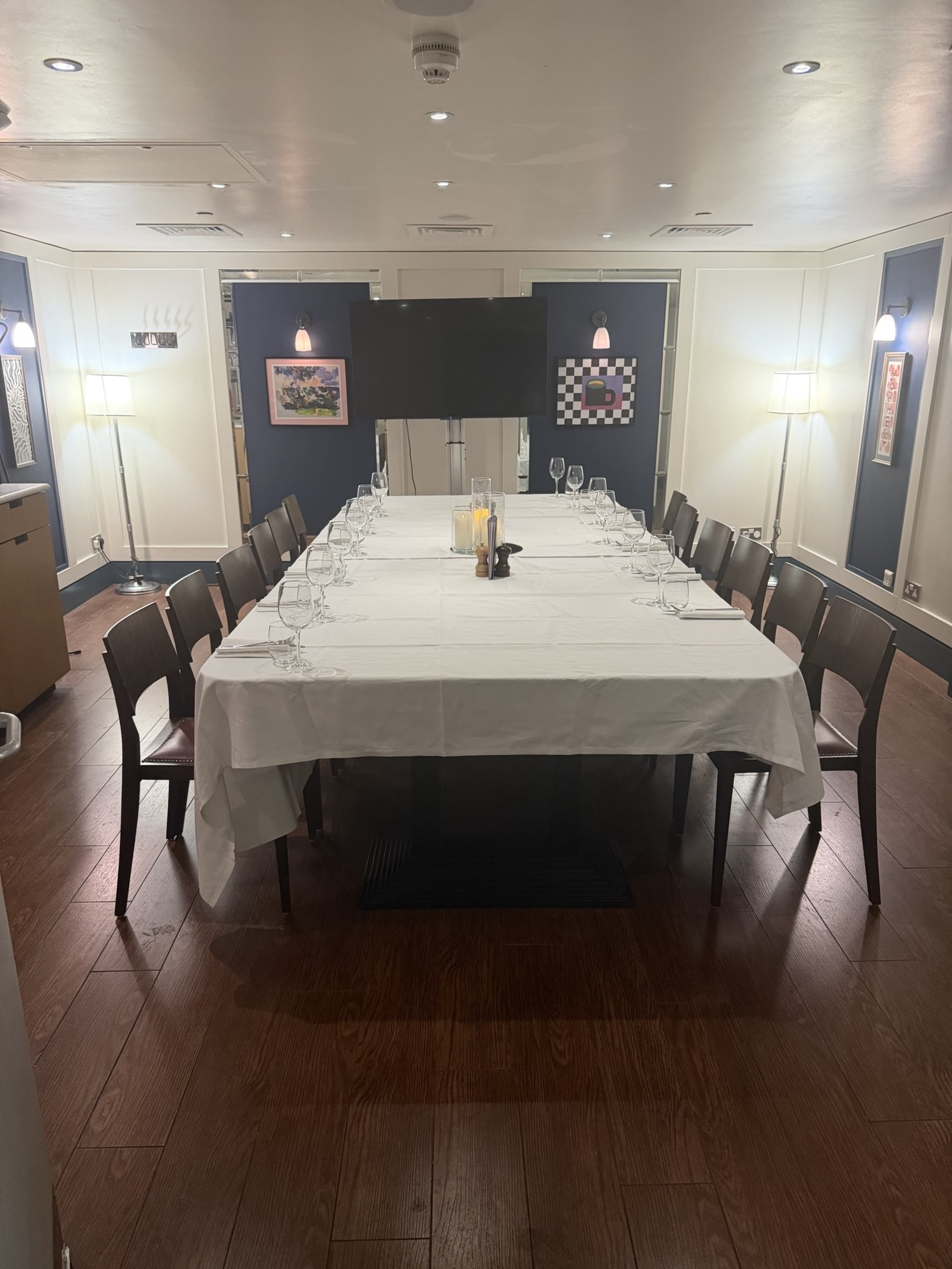 A long table is set for a formal dining arrangement under soft lighting in a room with artwork on the walls.