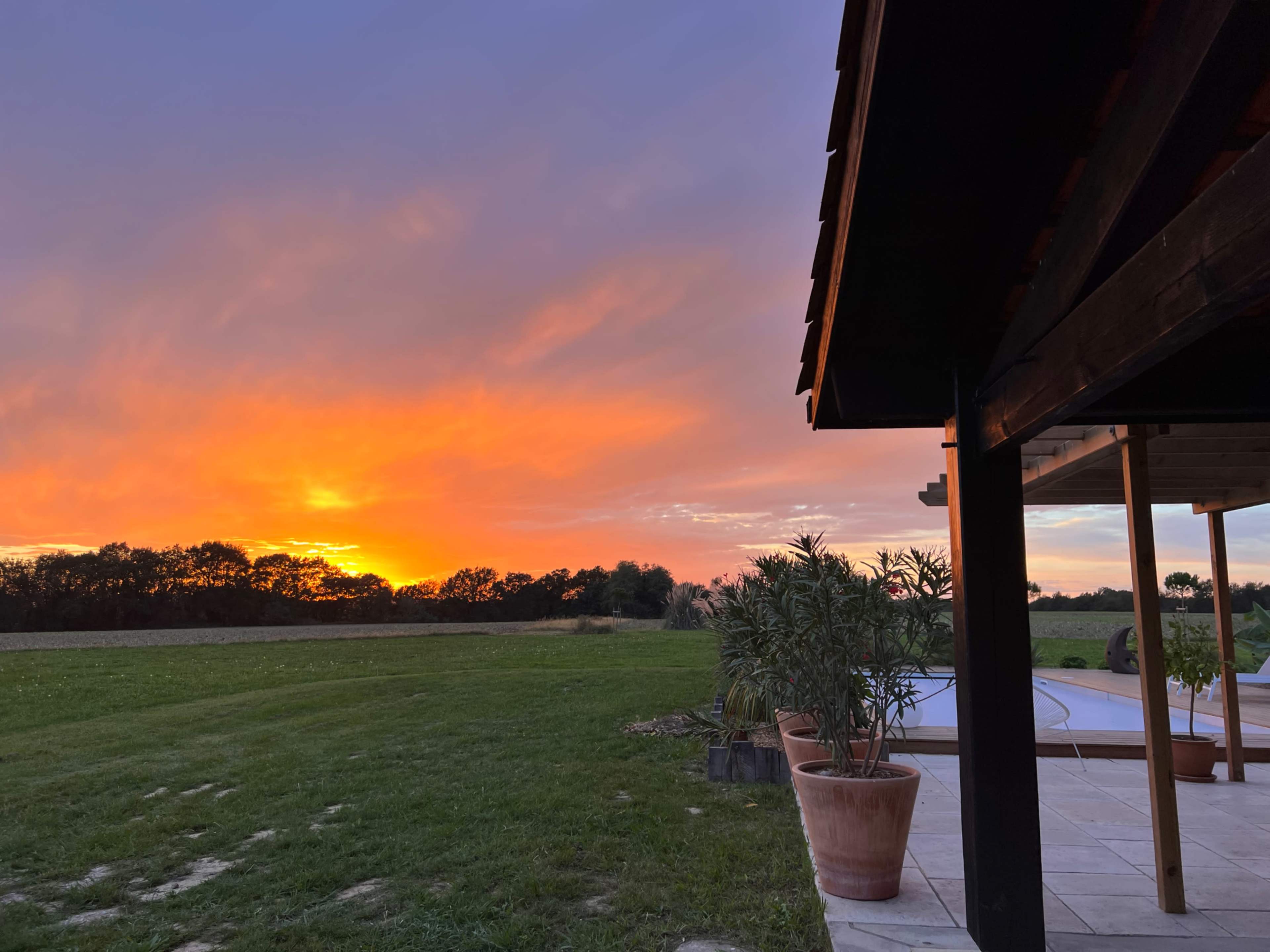 The scene shows a sunset with vibrant colors over a grassy landscape, framed by the corner of a building and potted plants.