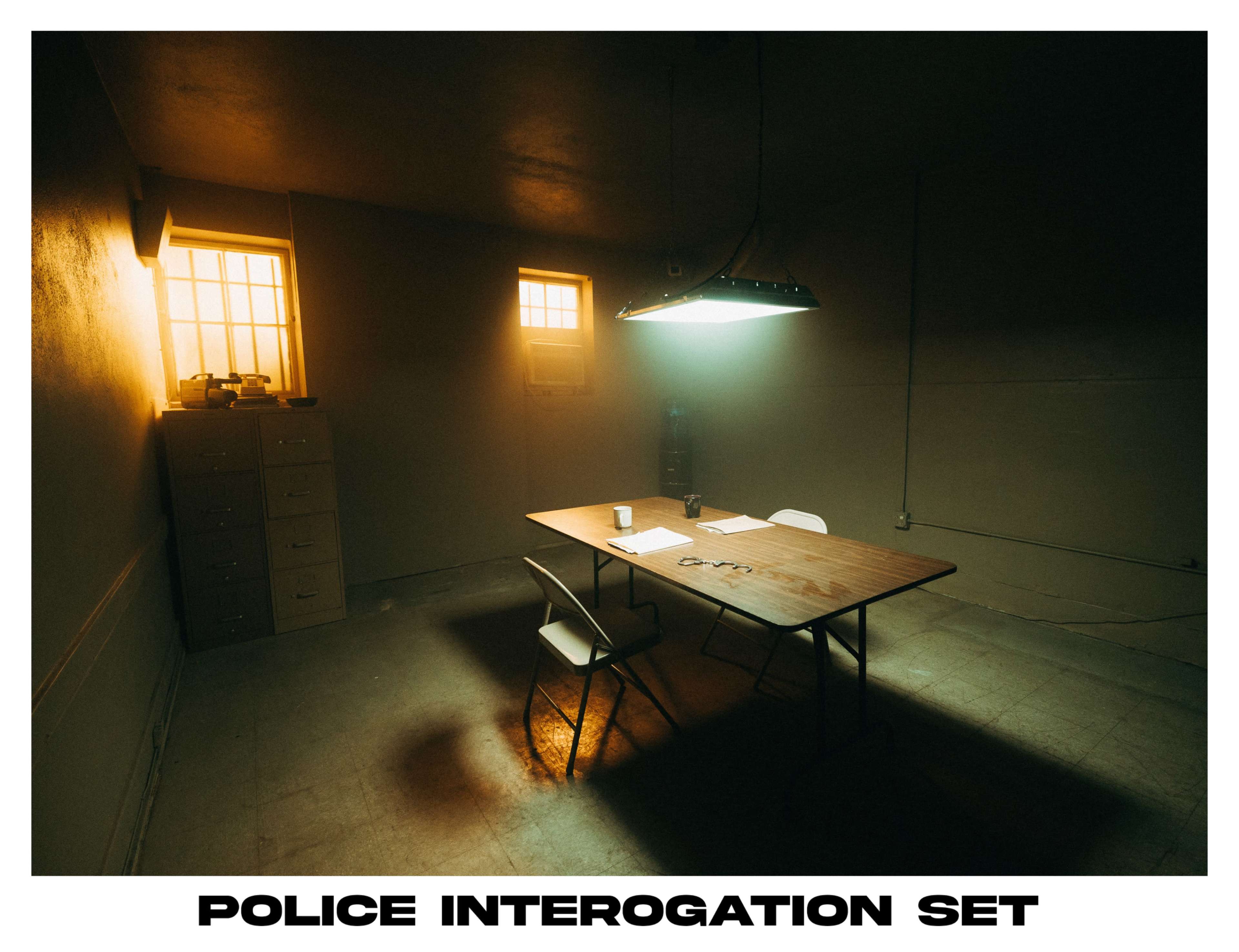A sparsely furnished police interrogation room features a table with chairs, dim lighting, and two small windows.
