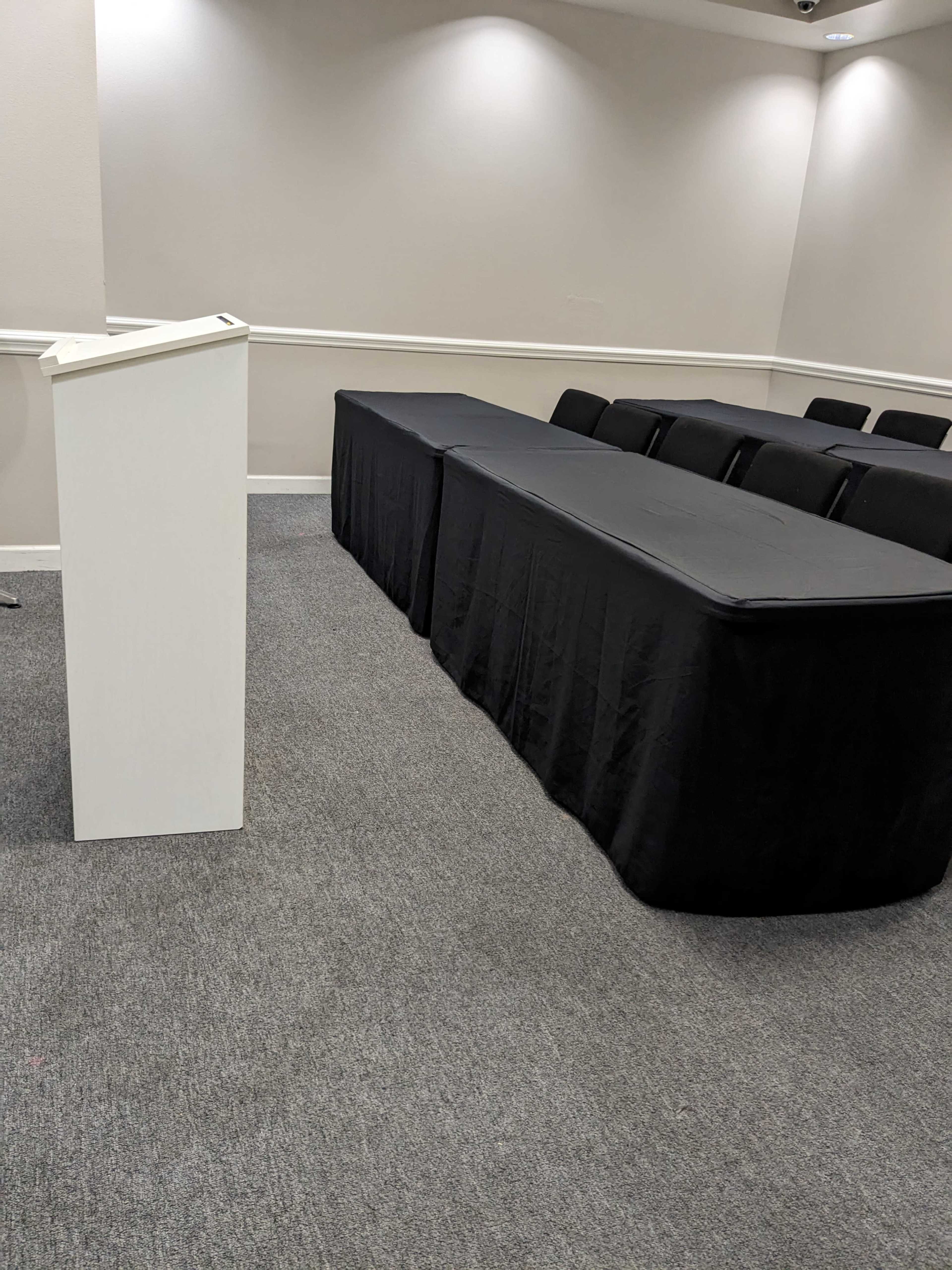 A small meeting room with a podium and a long black table covered with a cloth, accompanied by several chairs arranged in rows.