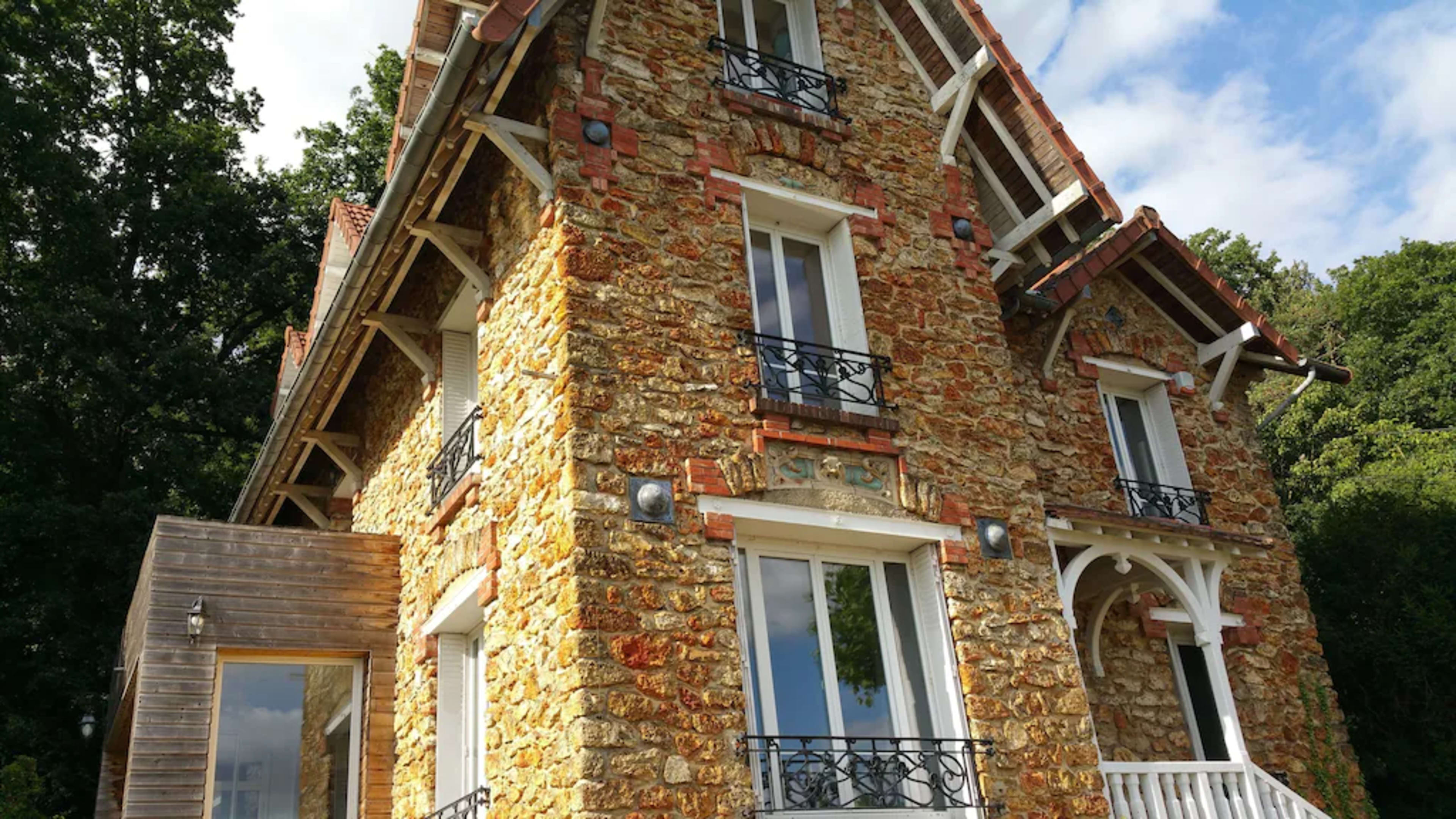 A three-story stone house with a prominent balcony and large windows, surrounded by trees.