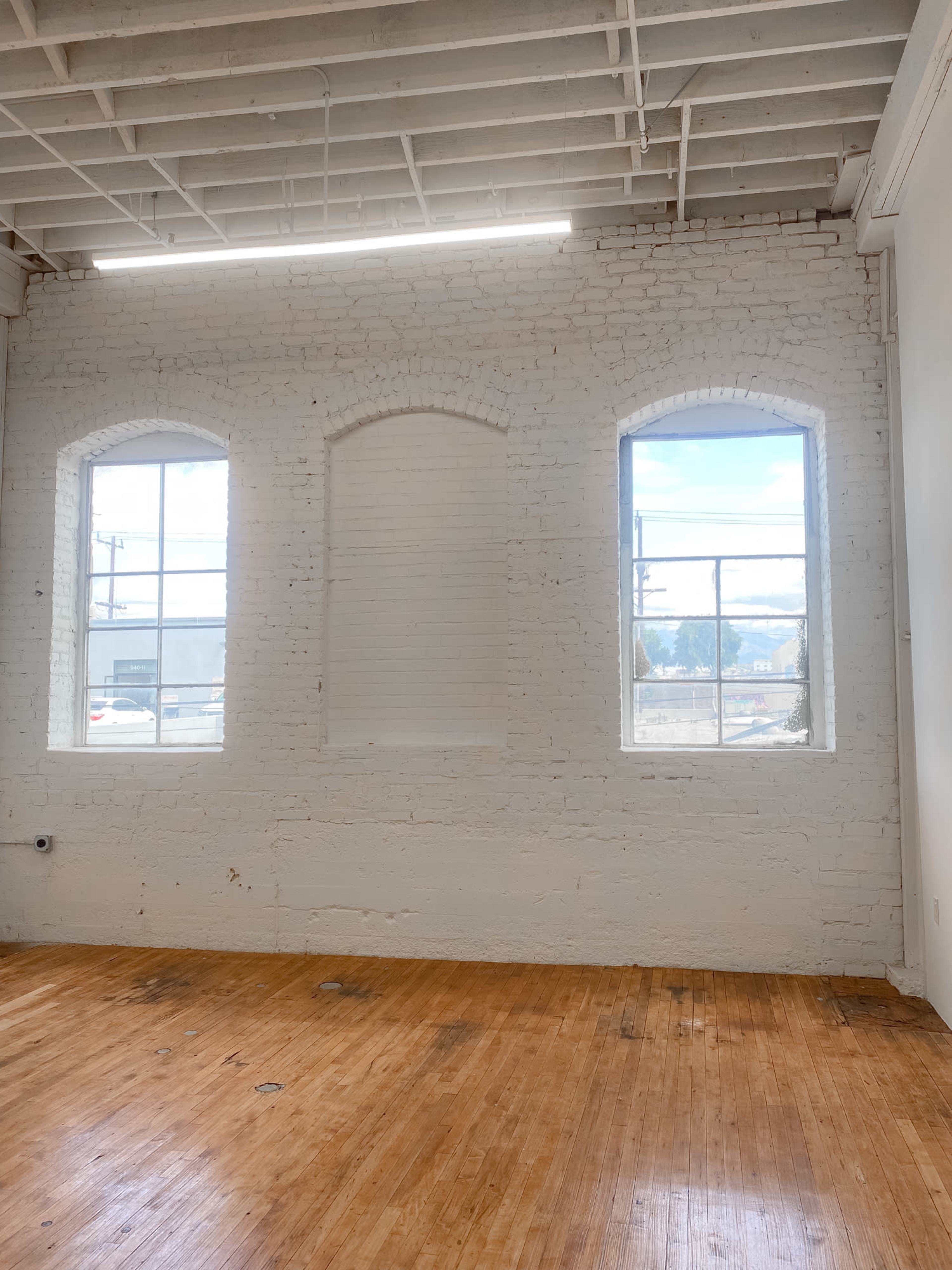 An empty room with three arched windows and wooden flooring, featuring a white brick wall.