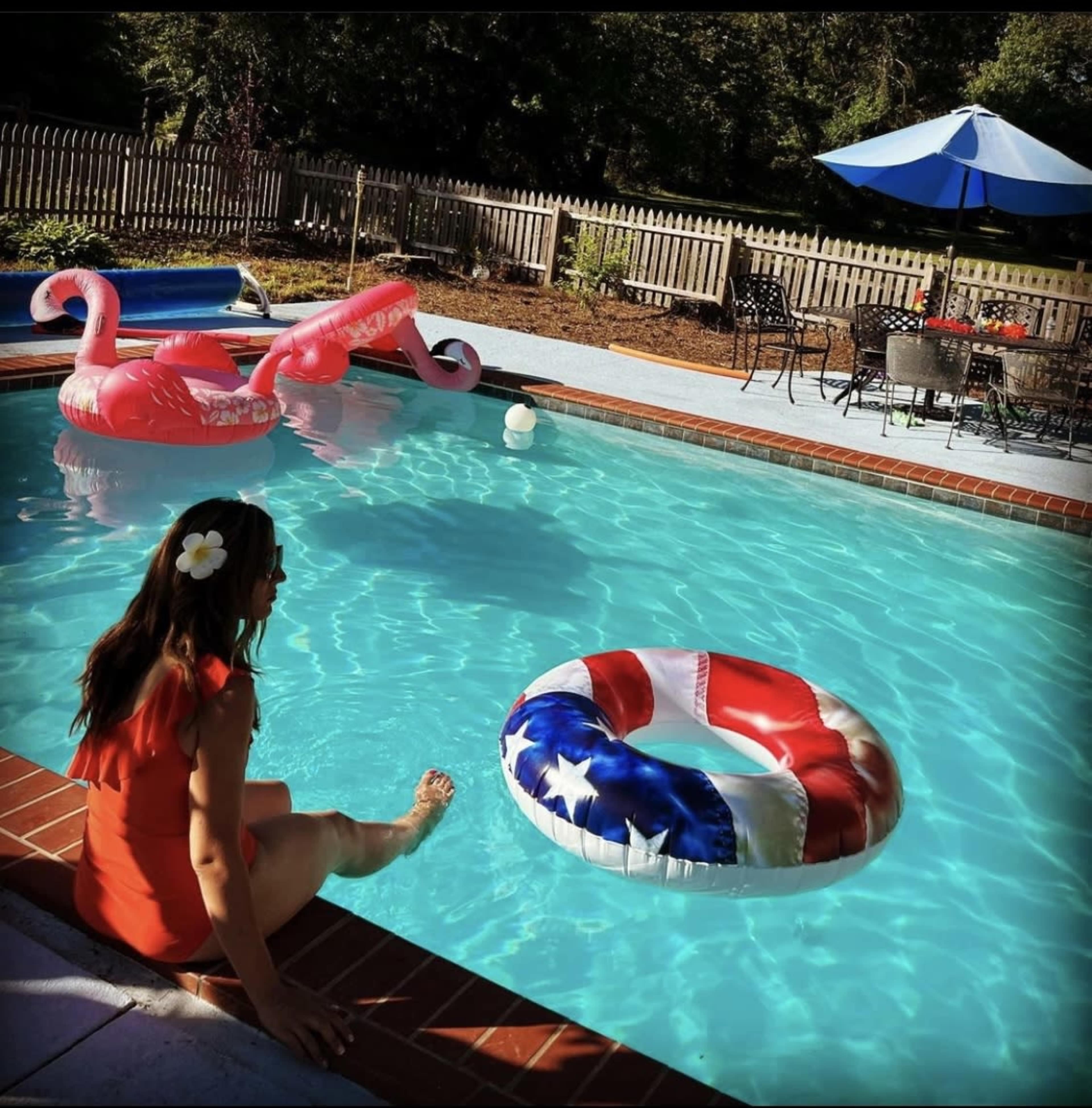 A person in a red dress sits at the edge of a pool, dipping their foot in the water beside colorful inflatable floats.