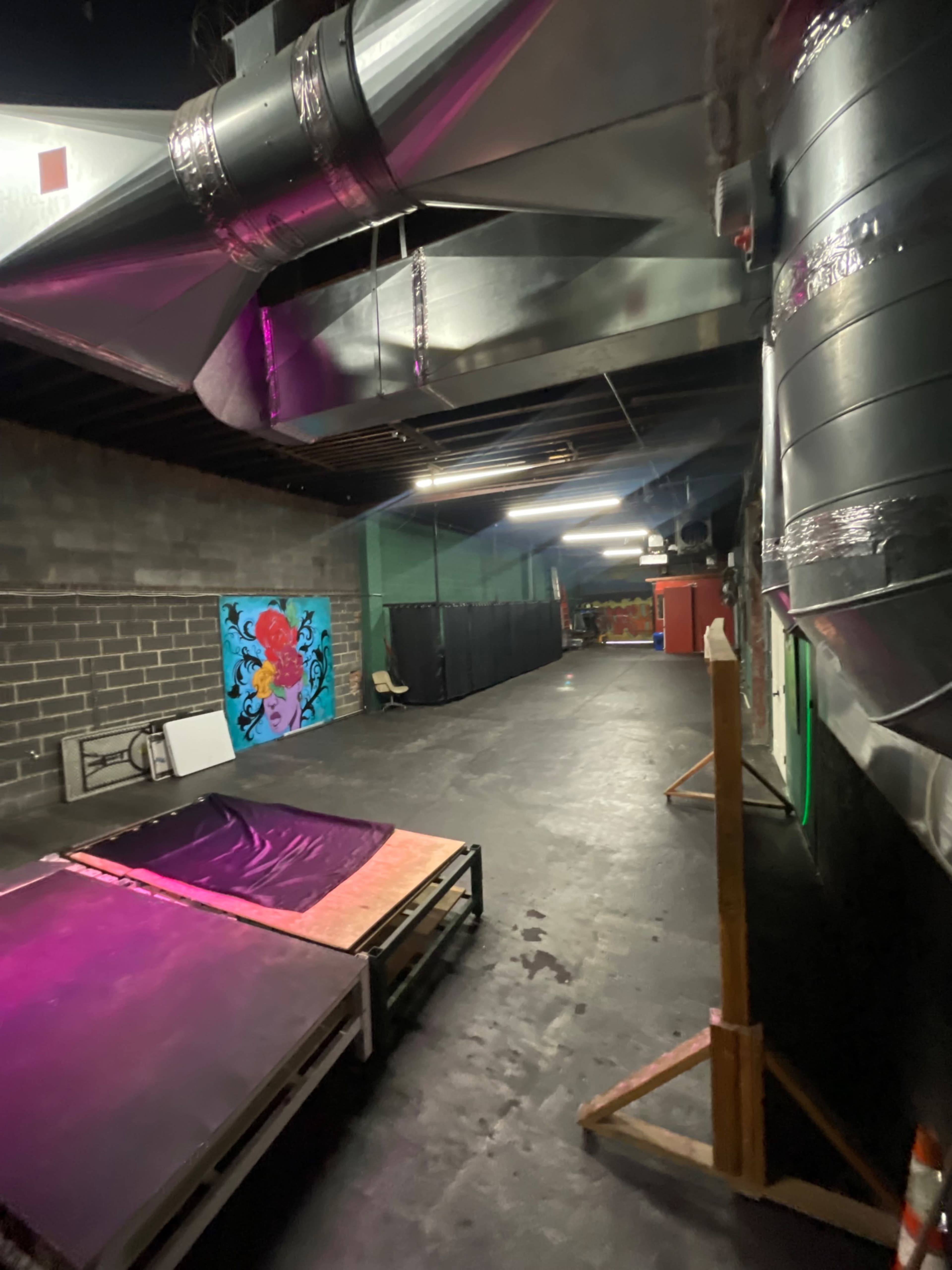 The image shows an industrial-style interior space with metal ductwork, colorful wall art, and two beds made from pallets on the floor.