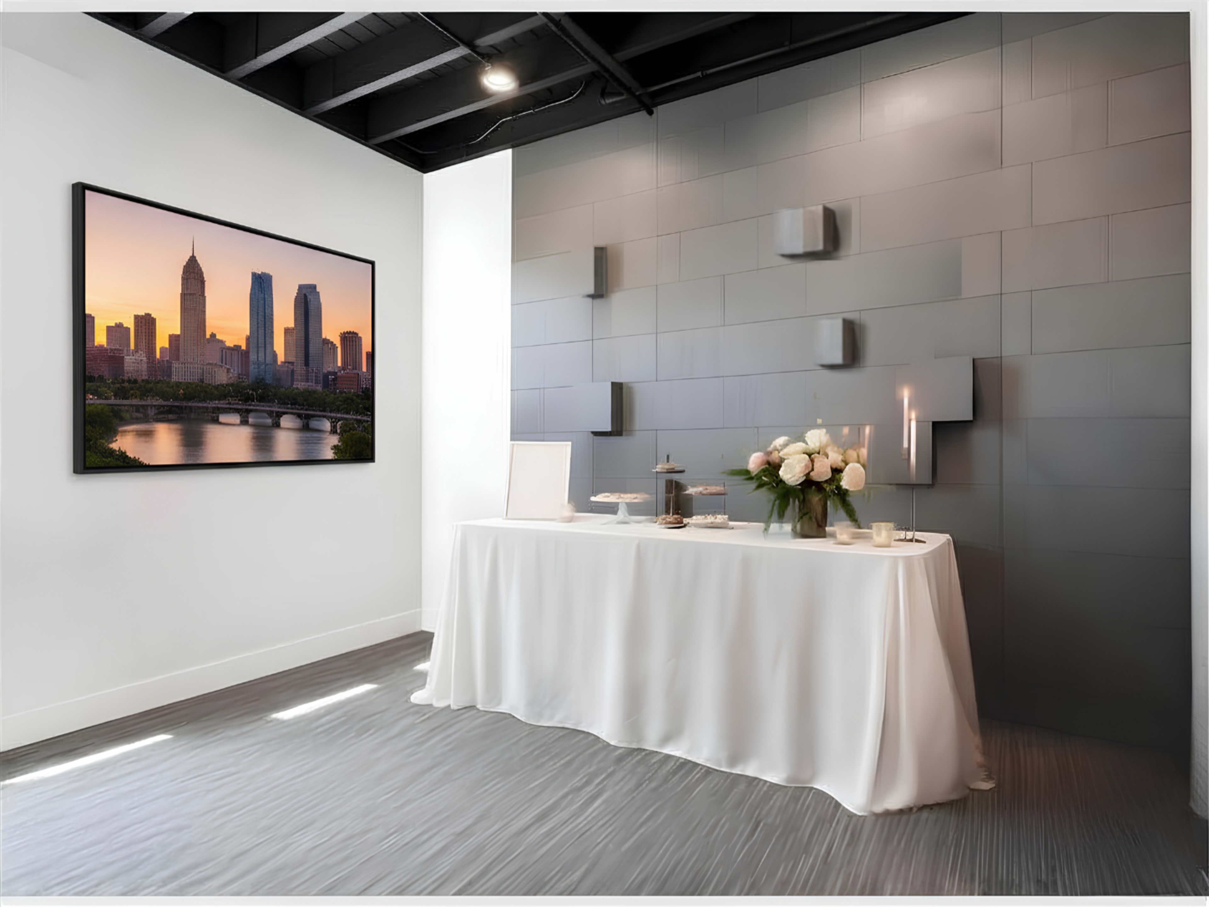 The image shows a modern event space with a white table covered in a cloth, adorned with flowers and candles, and a large framed cityscape photograph on the wall.