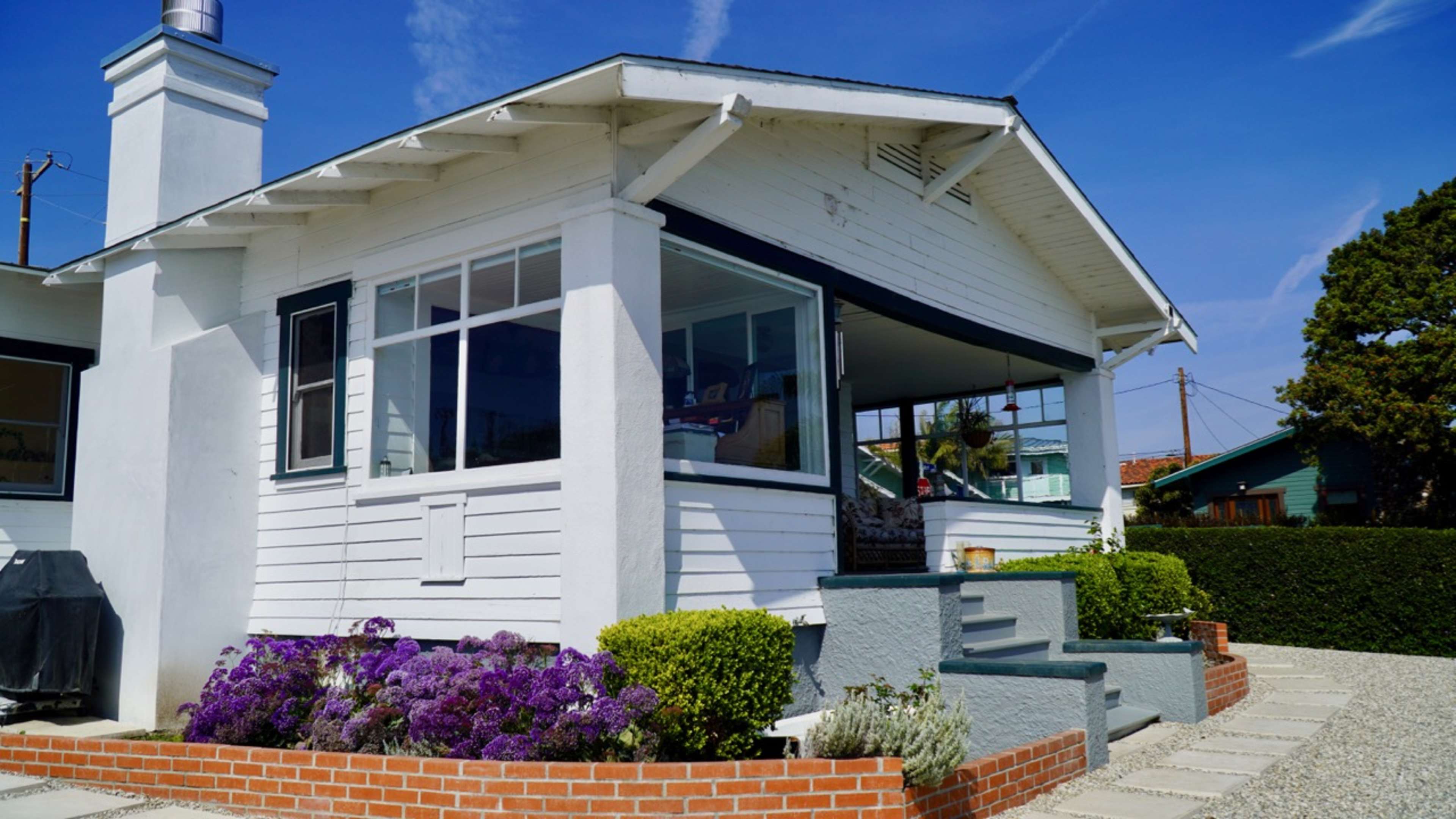 A white house with a covered porch features large windows and is surrounded by purple flowers and neatly arranged stone pathways.