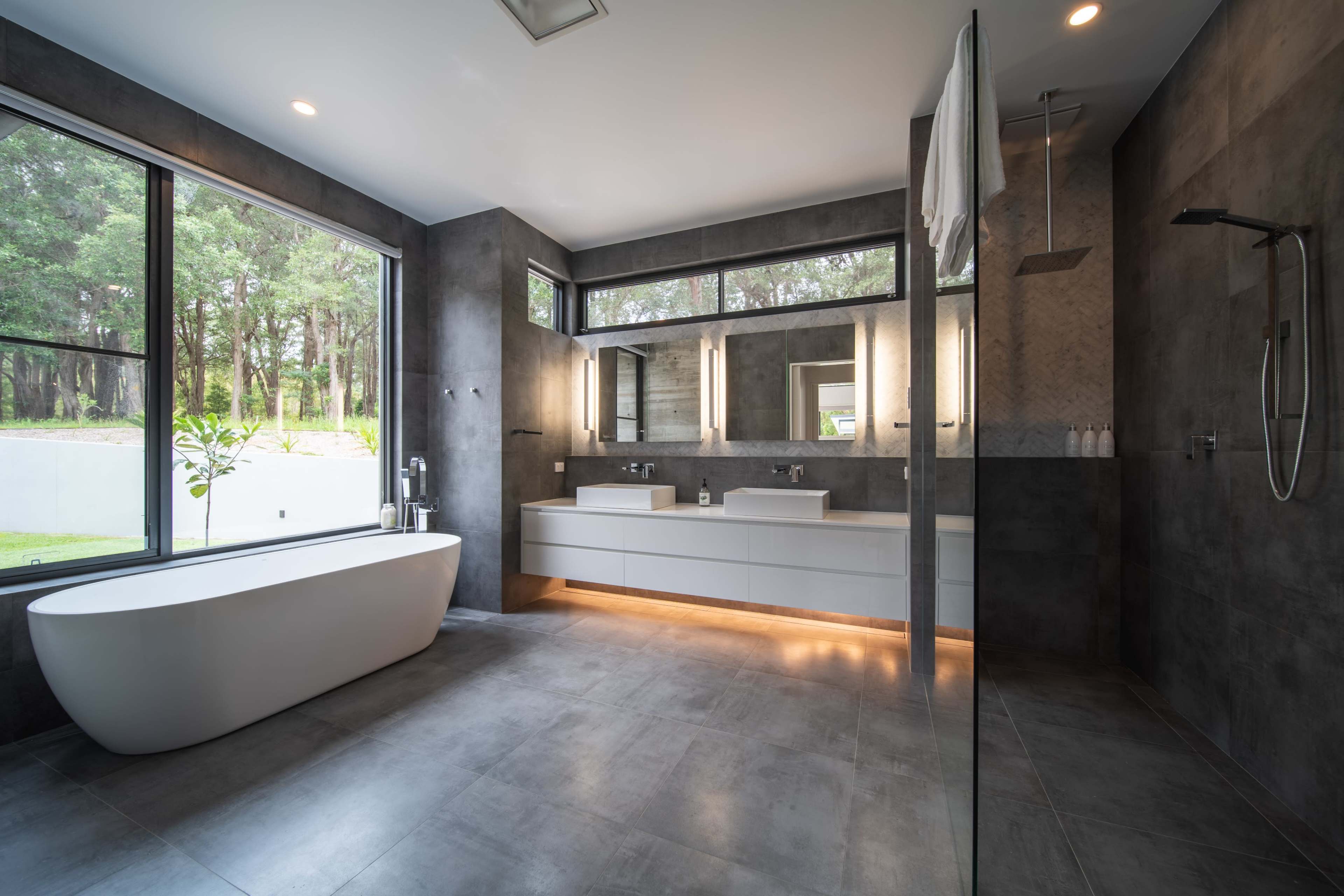 A modern bathroom featuring a freestanding bathtub, dual sinks with ample counter space, and large windows overlooking greenery.