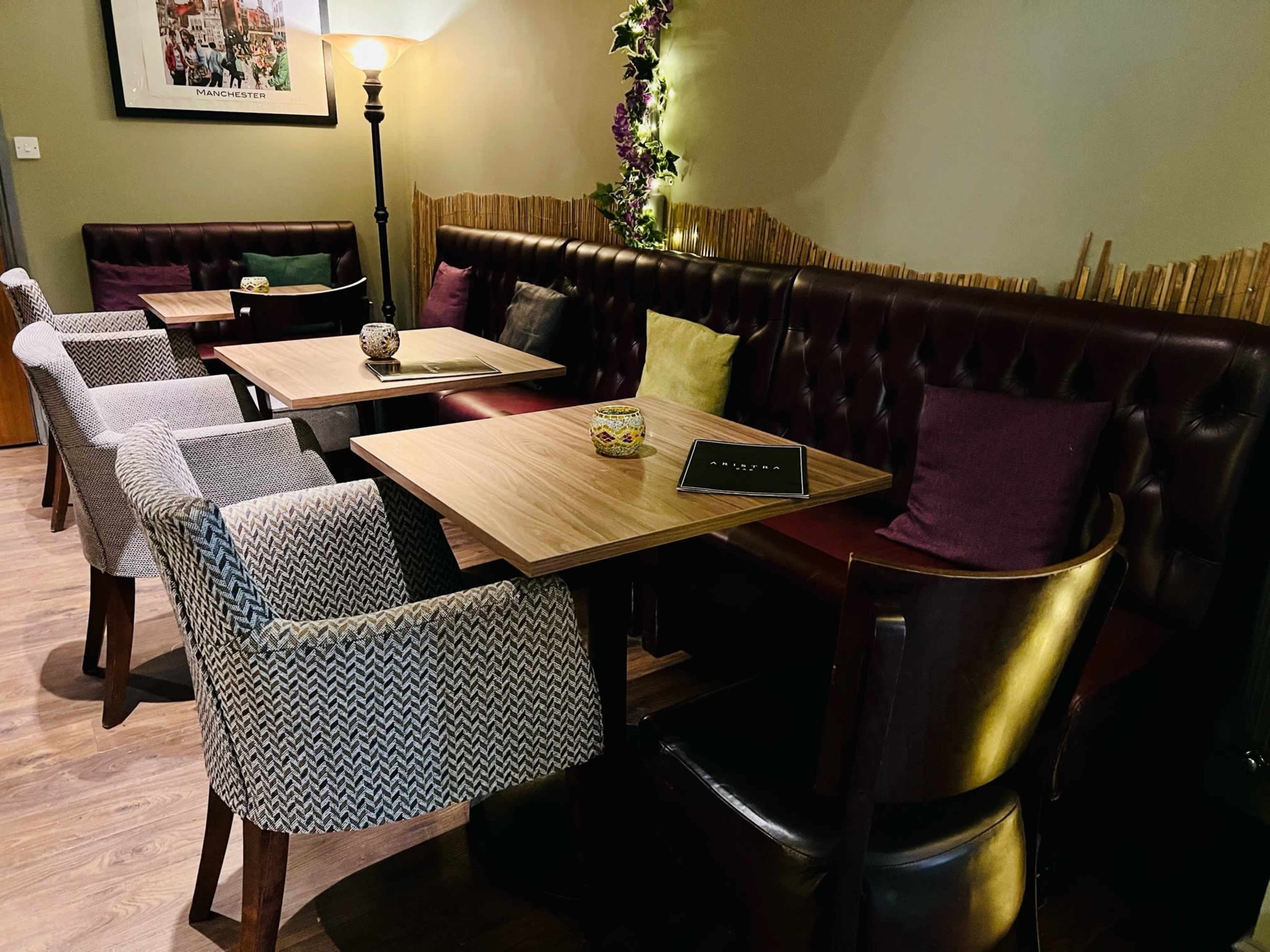 The image shows a cozy seating area in a dimly lit space, featuring several tables surrounded by upholstered chairs and a decorative lamp.