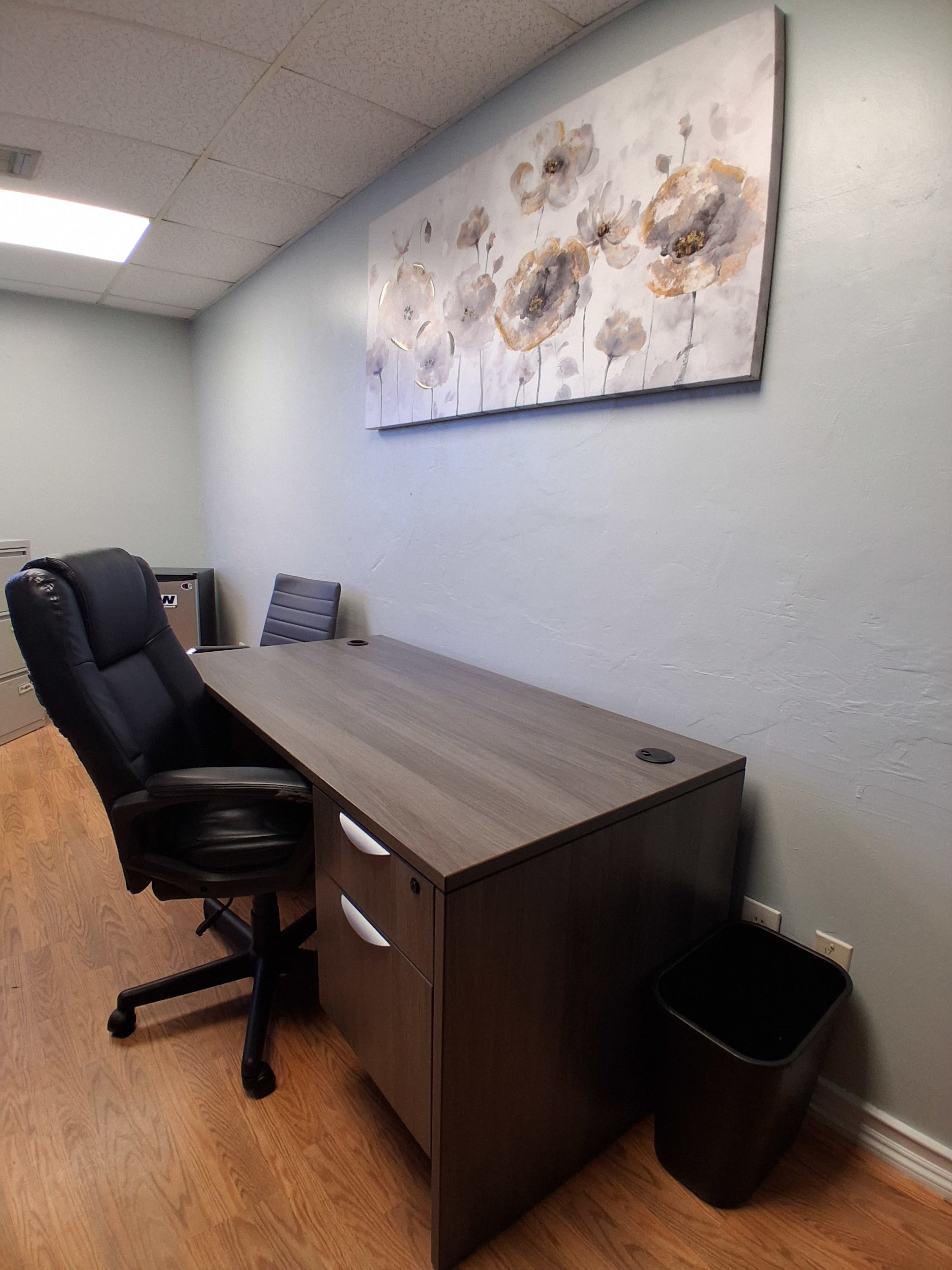 The image shows a minimalist office space featuring a desk with a chair, a black trash can, and a floral artwork on the wall.