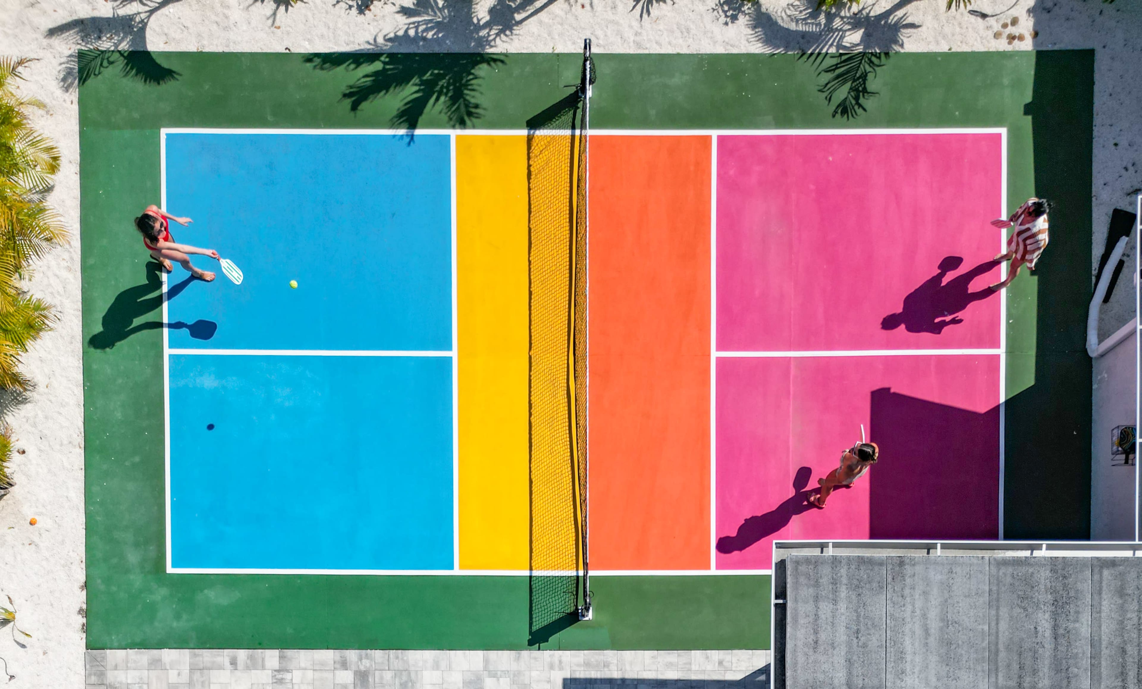 Two people are playing tennis on a brightly colored court divided into four sections of pink, yellow, blue, and green.