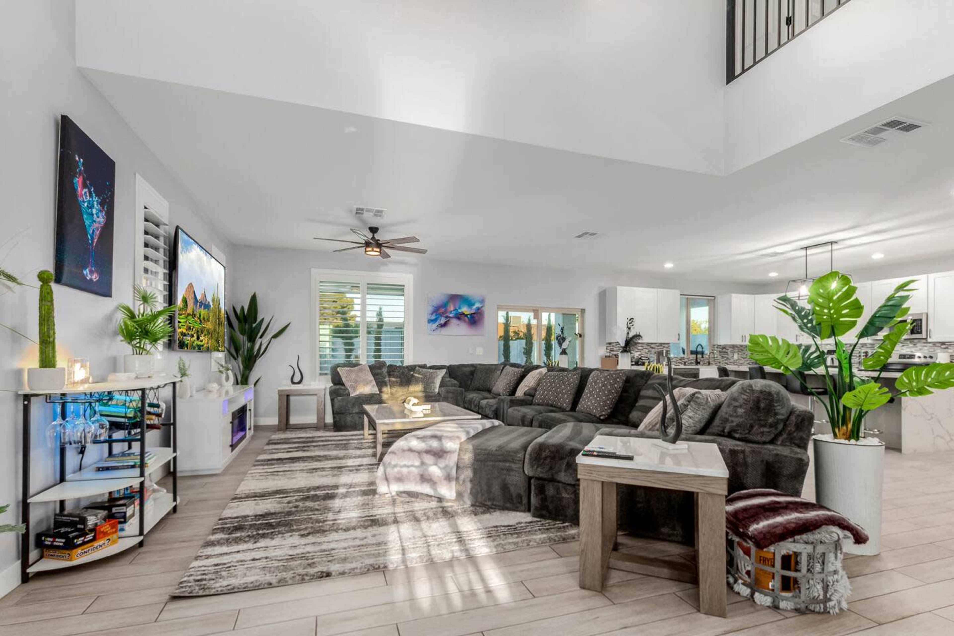 The image shows a spacious modern living room featuring a large sectional sofa, decorative plants, a coffee table, and an open layout leading to a dining area and kitchen.