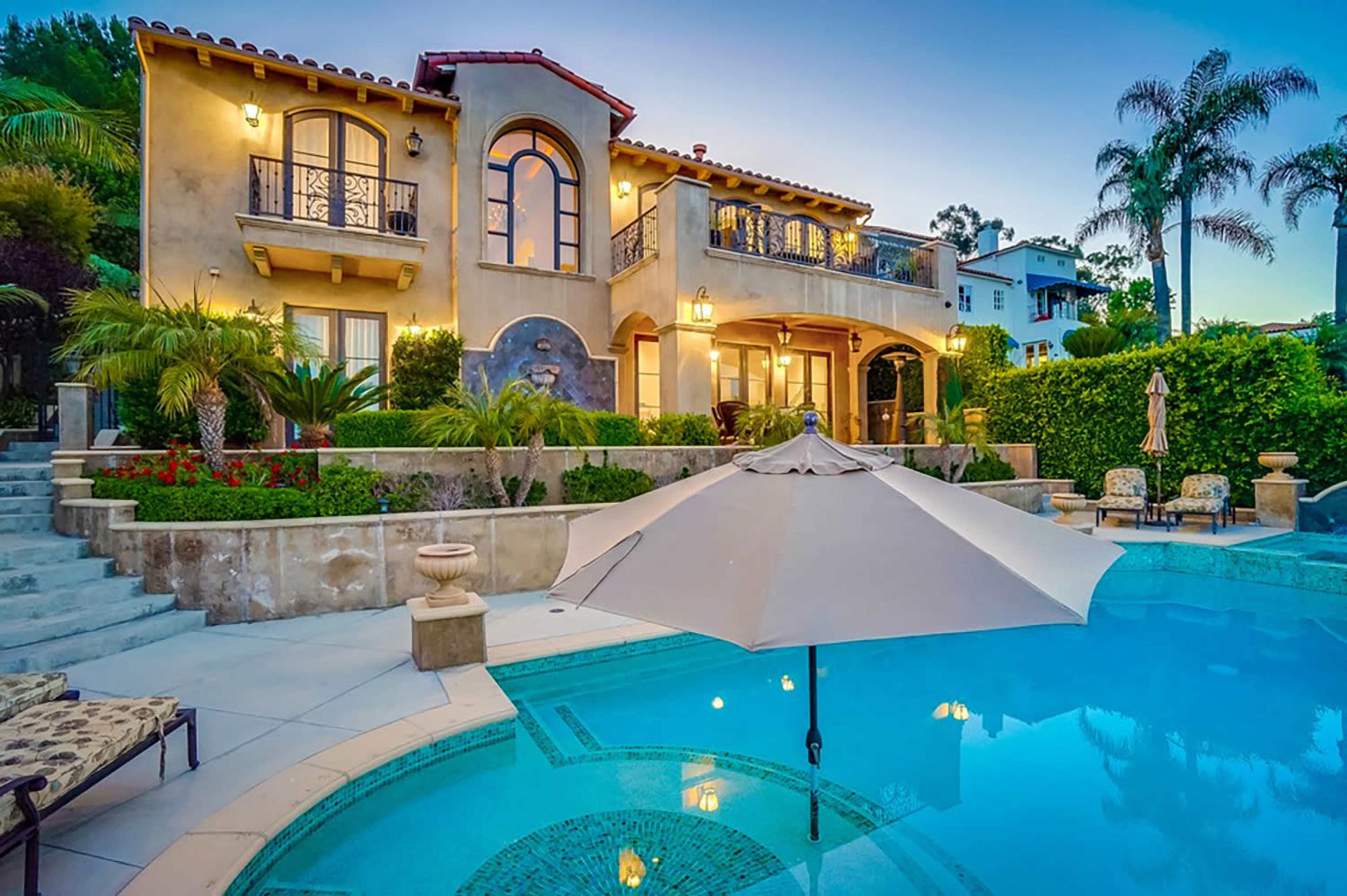 A beautifully designed two-story house with a pool and an umbrella in the foreground, surrounded by tropical plants and evening lighting.