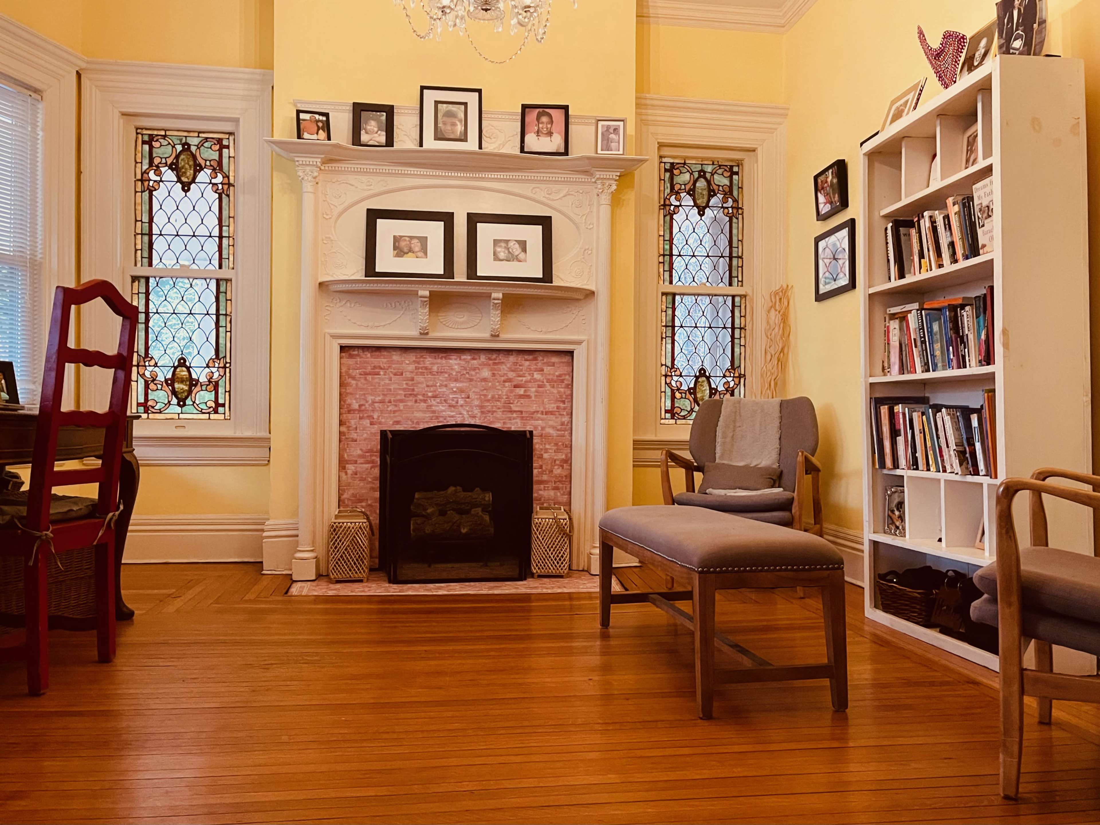 The room features a fireplace with a pink brick surround, framed artwork on the walls, a bookshelf, and several pieces of wooden furniture.